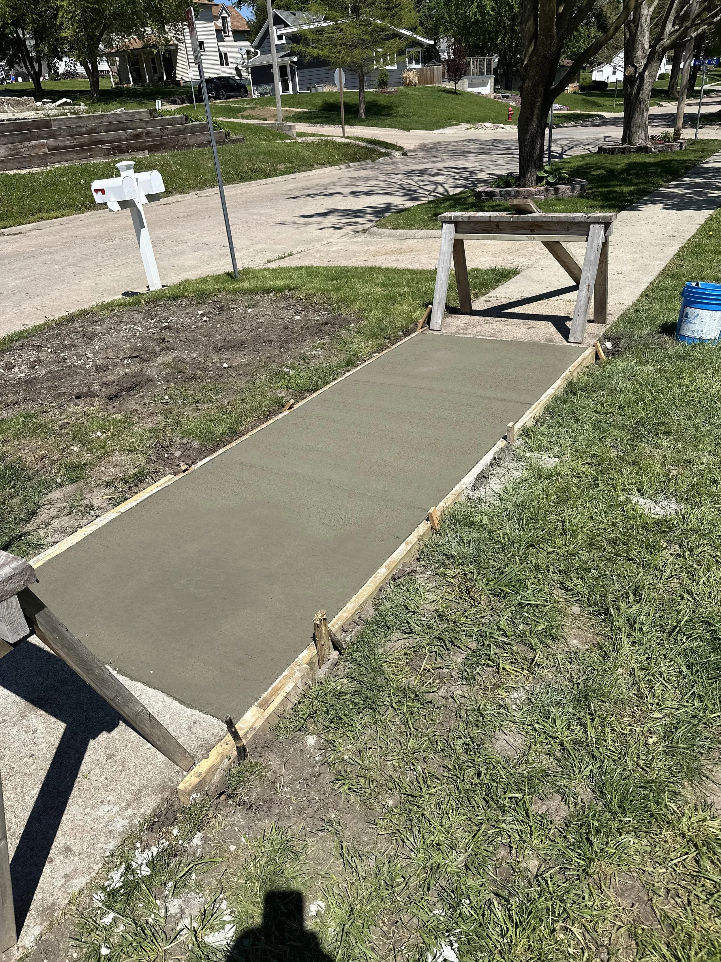 Newly poured concrete sidewalk being finished on a residential street, with wooden form still in place, and construction tools nearby.