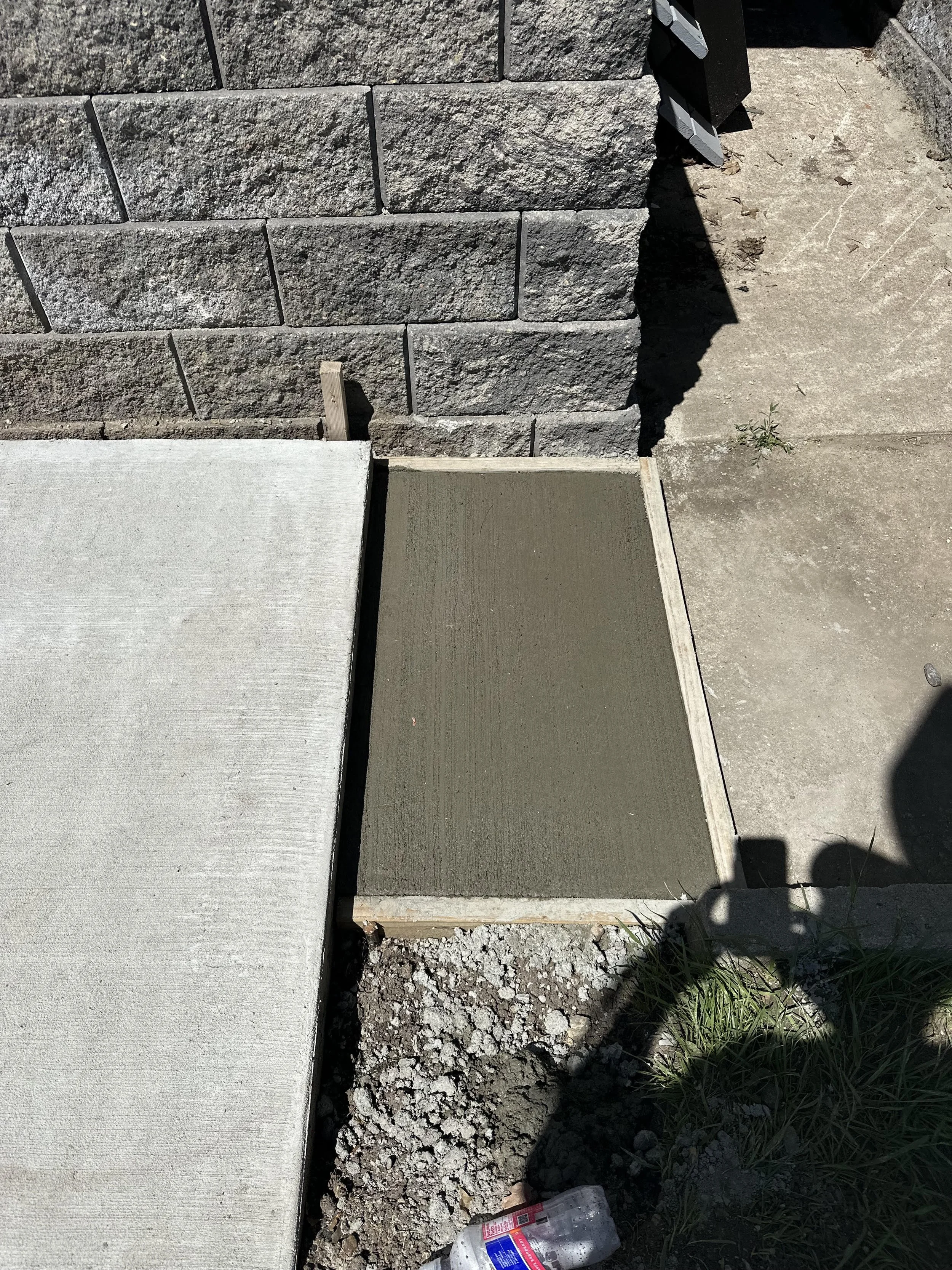 Newly poured concrete sidewalk section next to a brick wall and existing concrete path during construction.