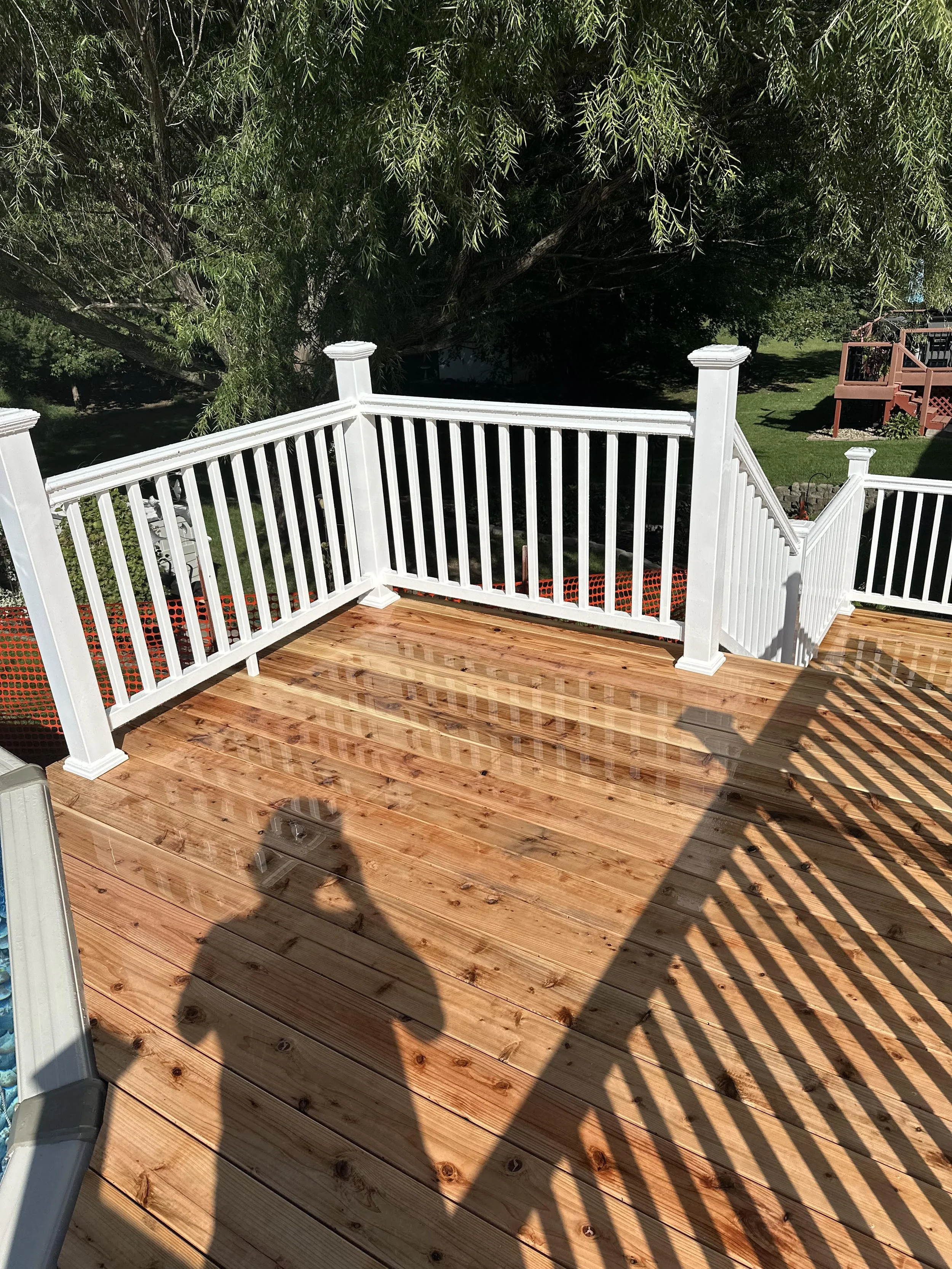 View of a newly built wooden deck with white railing, casting shadows on the wood floor, with green trees and a grassy yard in the background.