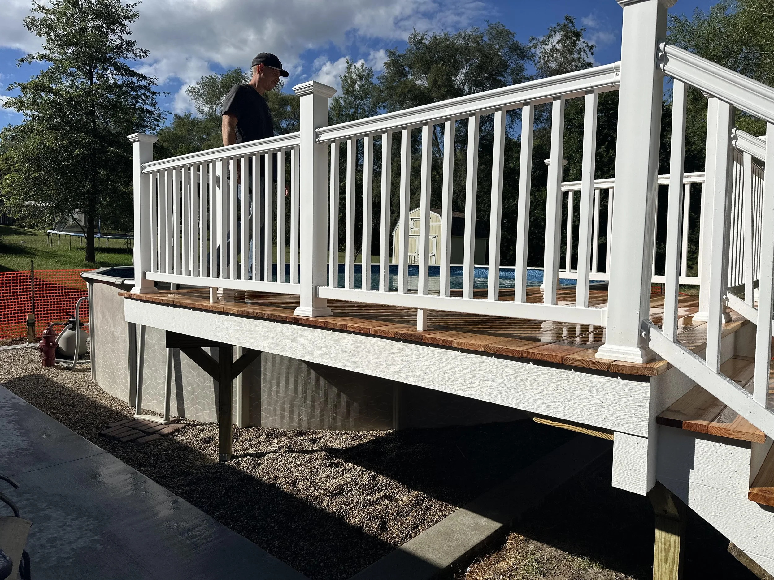 Person standing on a wooden deck with a white railing, overlooking a backyard with a pool and trees.