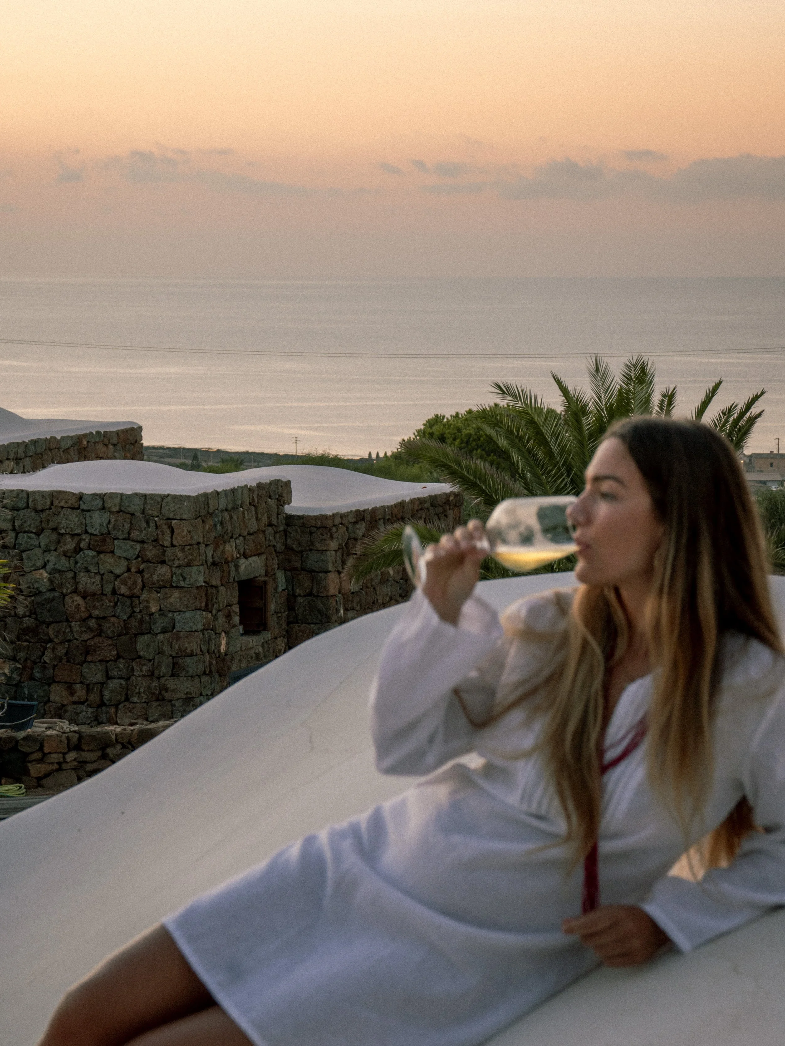 Una donna seduta sui tetti bianchi dei Dammusi che beve da un bicchiere di vino bianco, con un panorama di mare e cielo al tramonto in background.