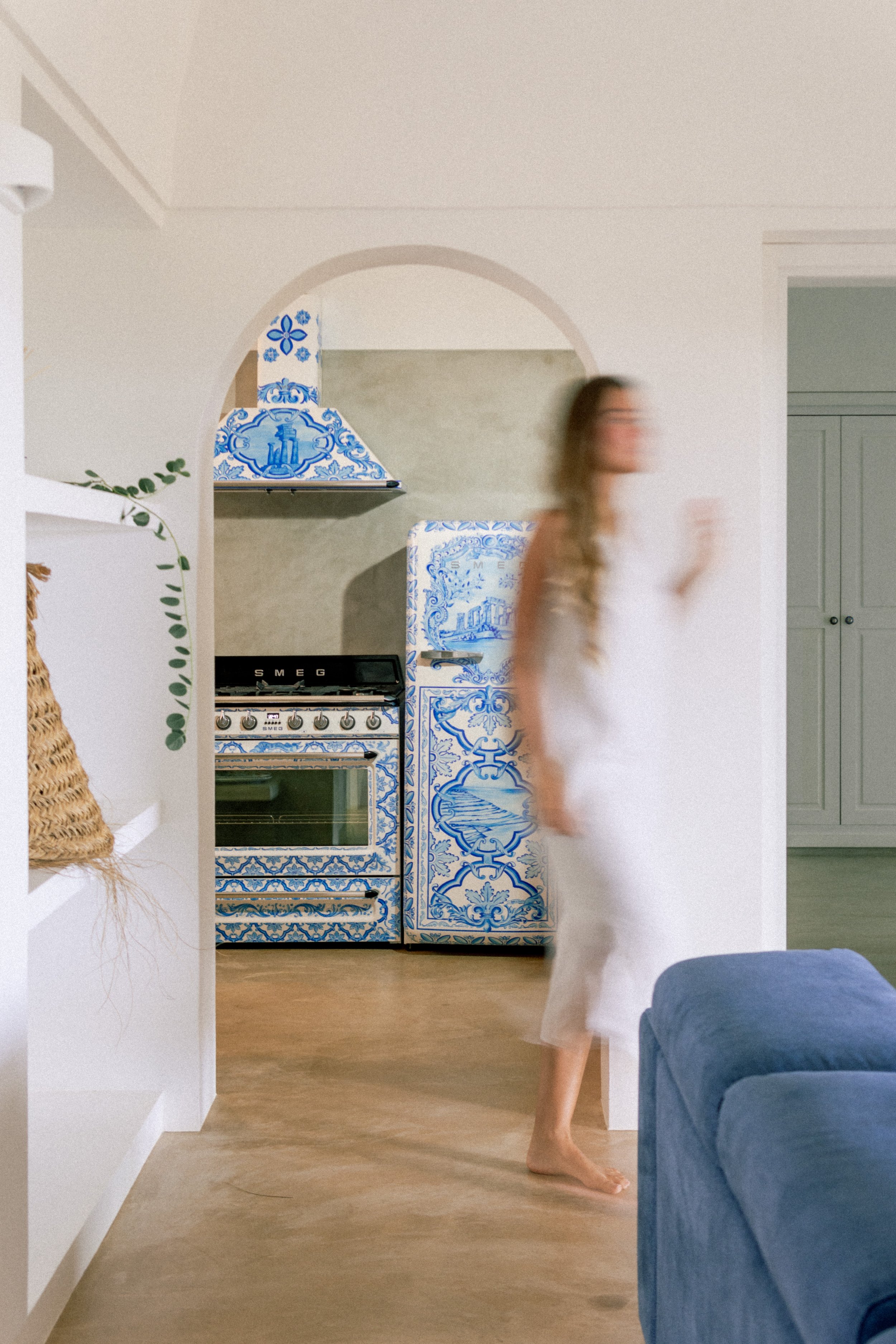 Cucina con elettrodomestici Dolce & Gabbana e una donna in un abito bianco che cammina attraverso un soggiorno con divano blu e scaffali con piante attorno.