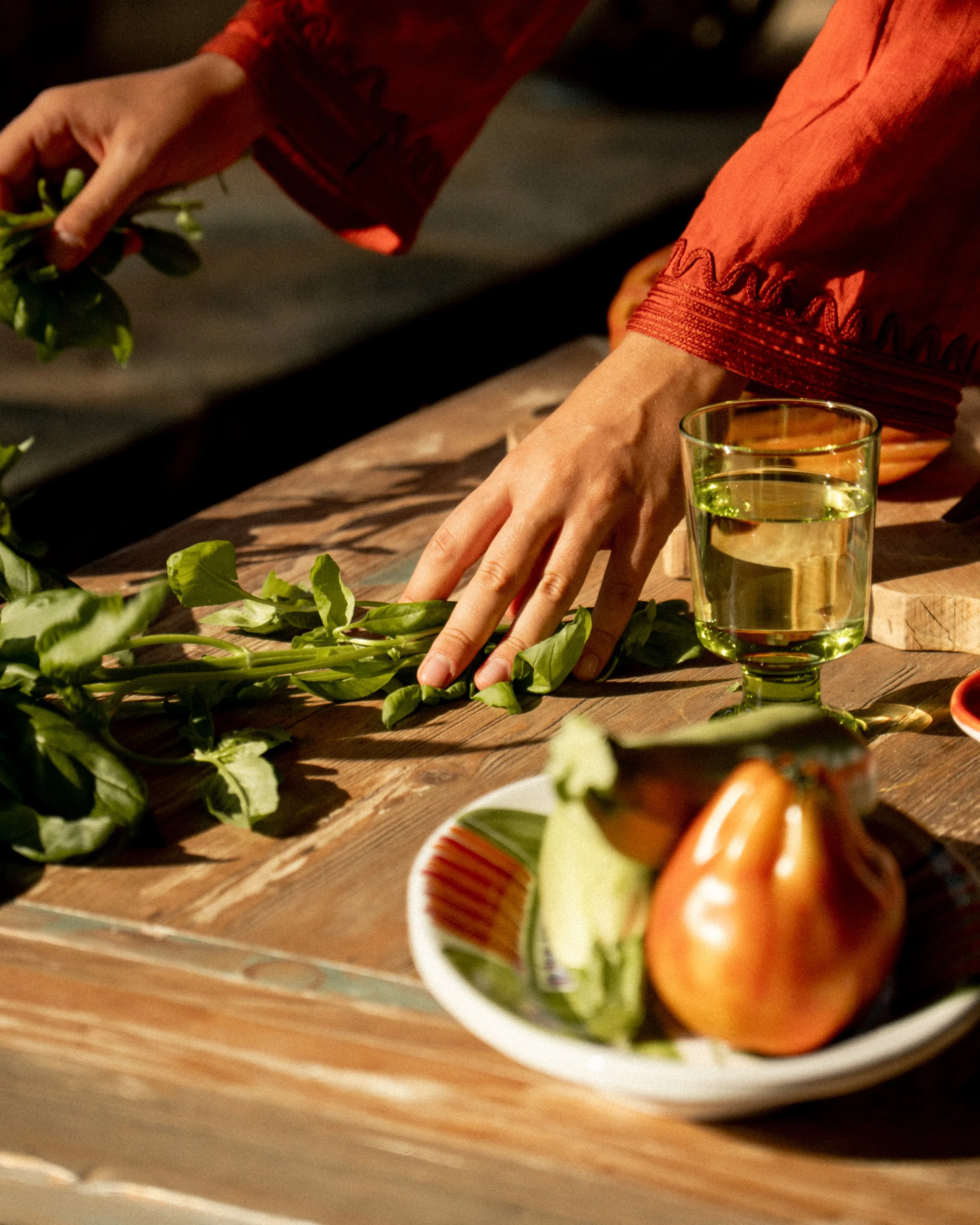 Una persona che sistema le foglie di basilico su un tavolo di legno, con un bicchiere di acqua e un piatto con un pomodoro e capperi