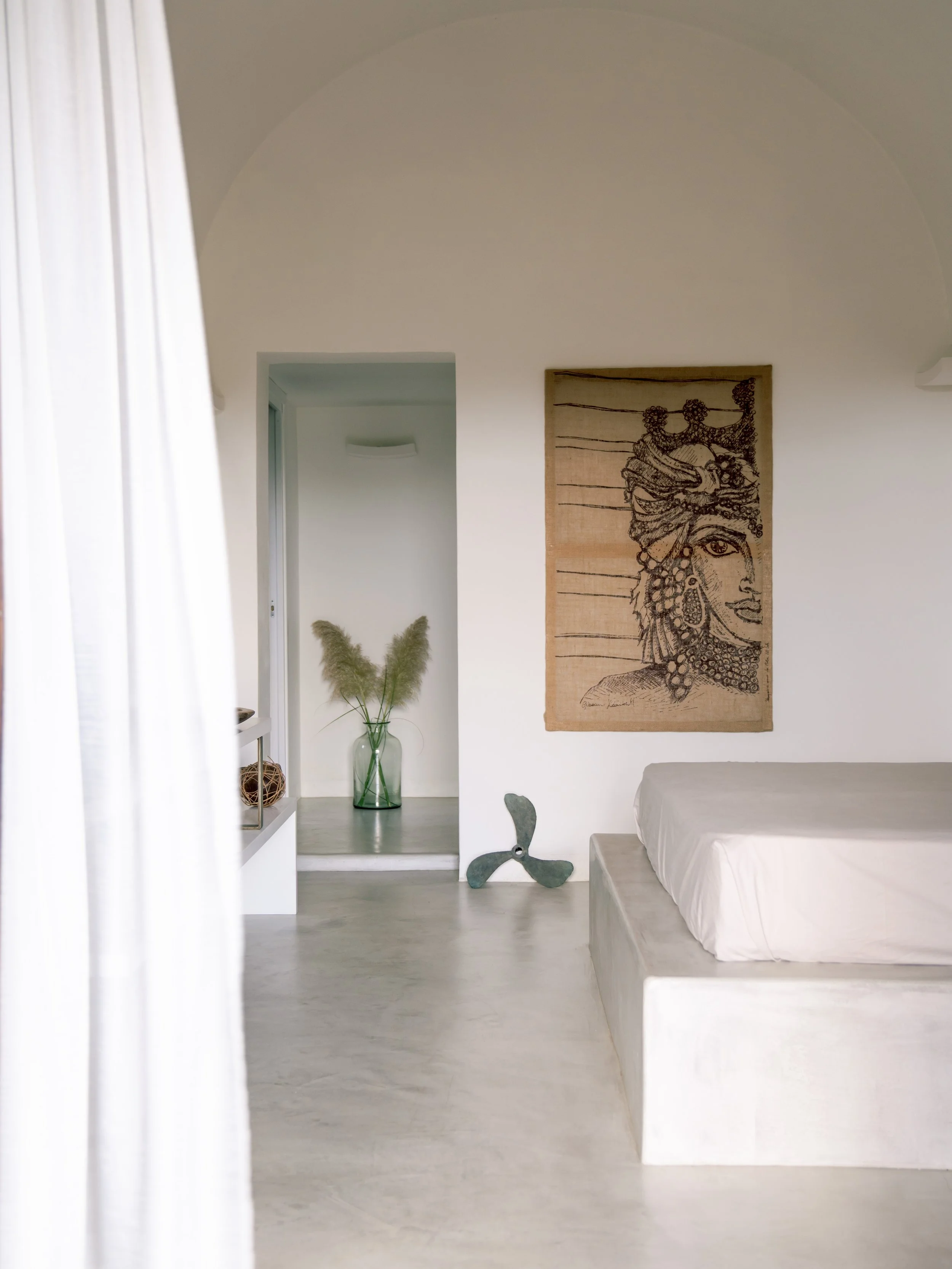 Interno di una stanza con pareti bianche, un letto con copripiumino bianco, un quadro con un volto di donna disegnata in stile etnico appeso alla parete, una decorazione con pale di grano in un vaso di vetro verde, una statuina di un elicottero in metallo e una porta aperta verso un piccolo ingresso con un vaso di fiori secchi.