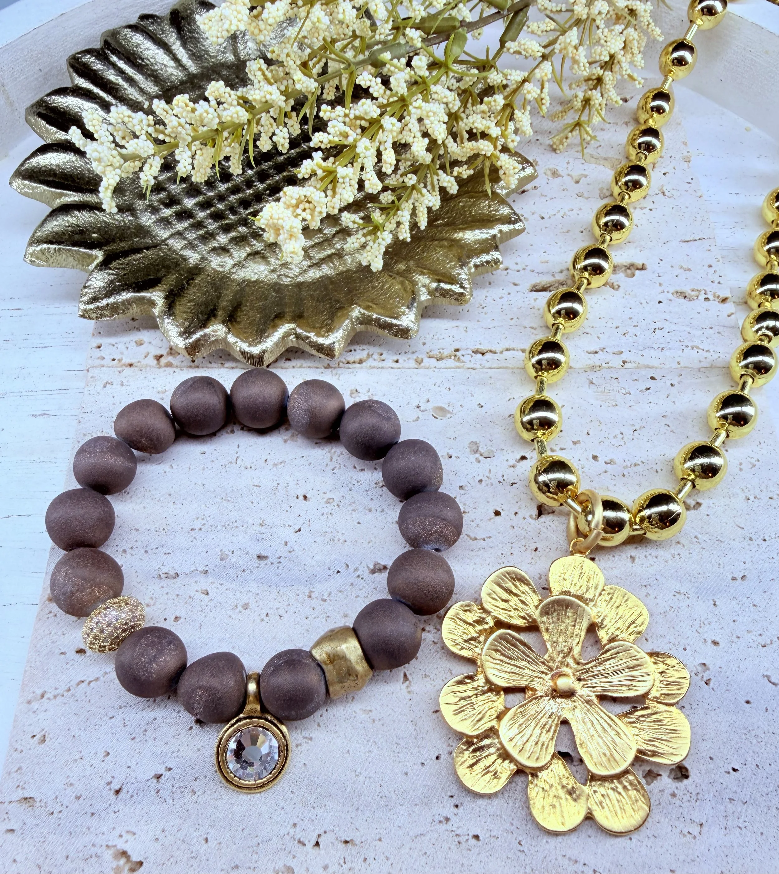 Gold and silver jewelry, including a large gold flower-shaped pendant, a black beaded necklace, a gold beaded necklace, and a crystal pendant, arranged with white flowers on a rustic white surface.