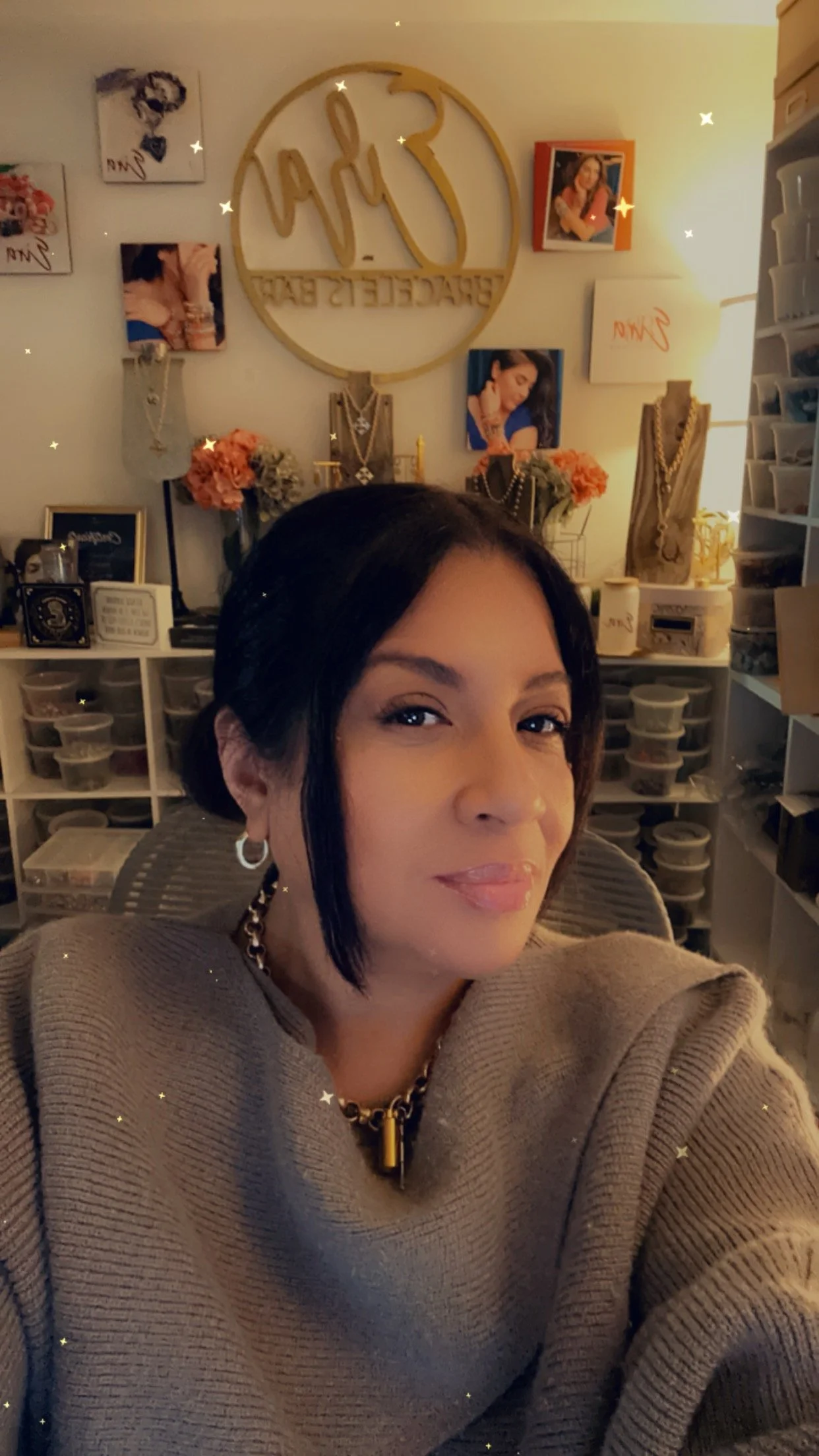 A woman with dark hair in a bob style, wearing a beige sweater and gold jewelry, takes a selfie in a room decorated with jewelry displays, artwork, and photos on the wall.