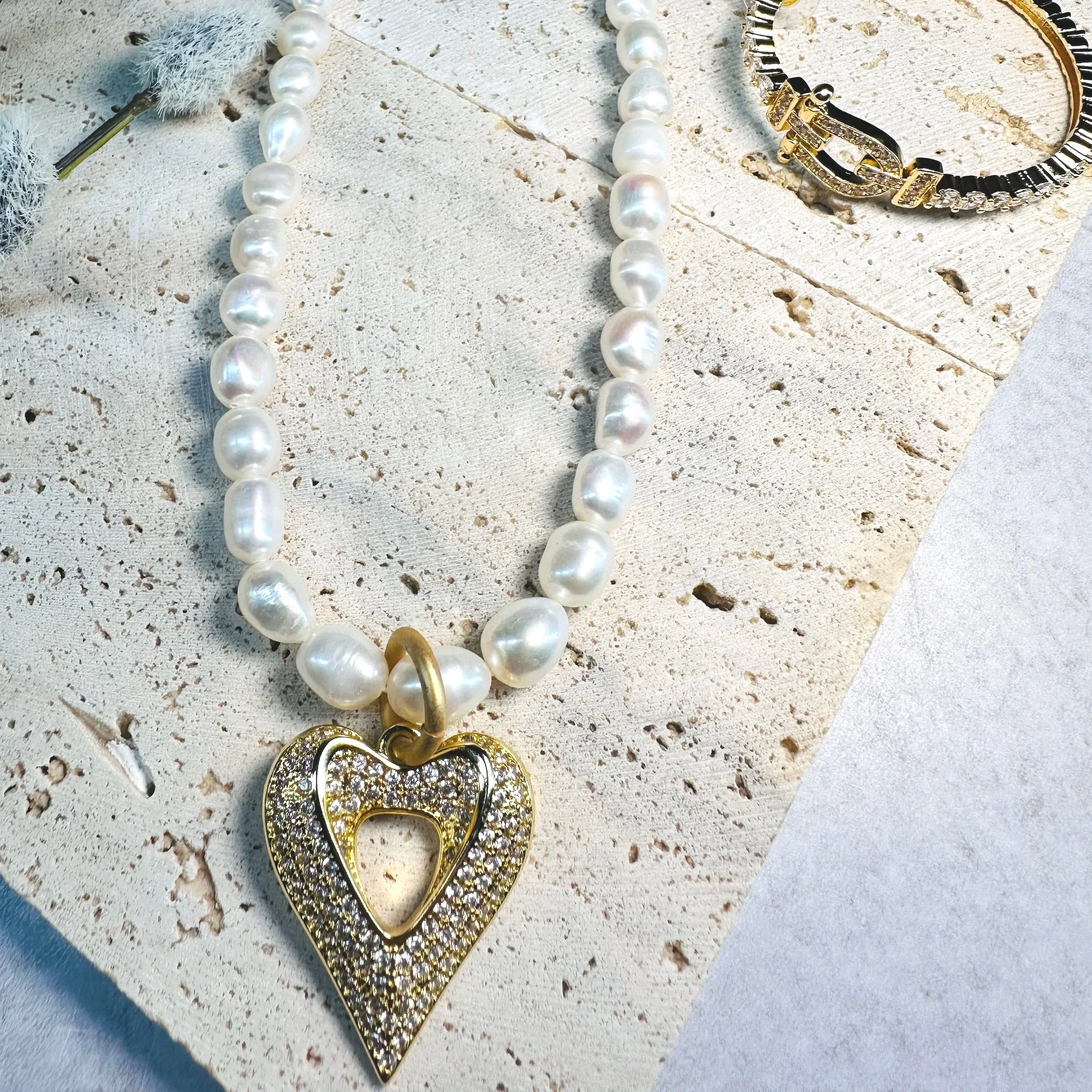 A pearl necklace with a gold and diamond heart pendant, a gold and diamond bracelet, and a silver and black bracelet, displayed on a textured white surface.