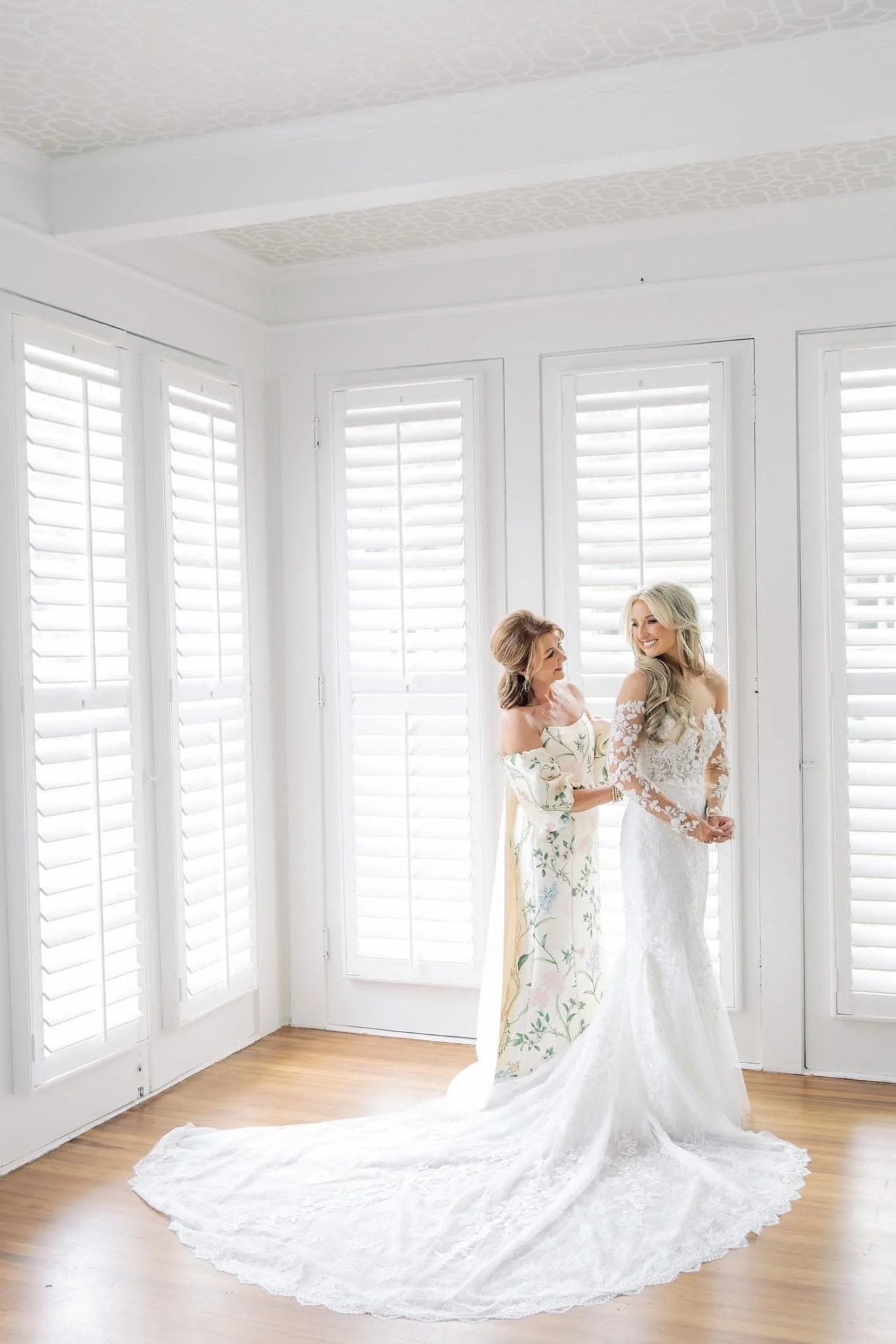 A bride in a wedding dress and an older woman in a floral dress, possibly her mother, standing in front of large white window shutters.