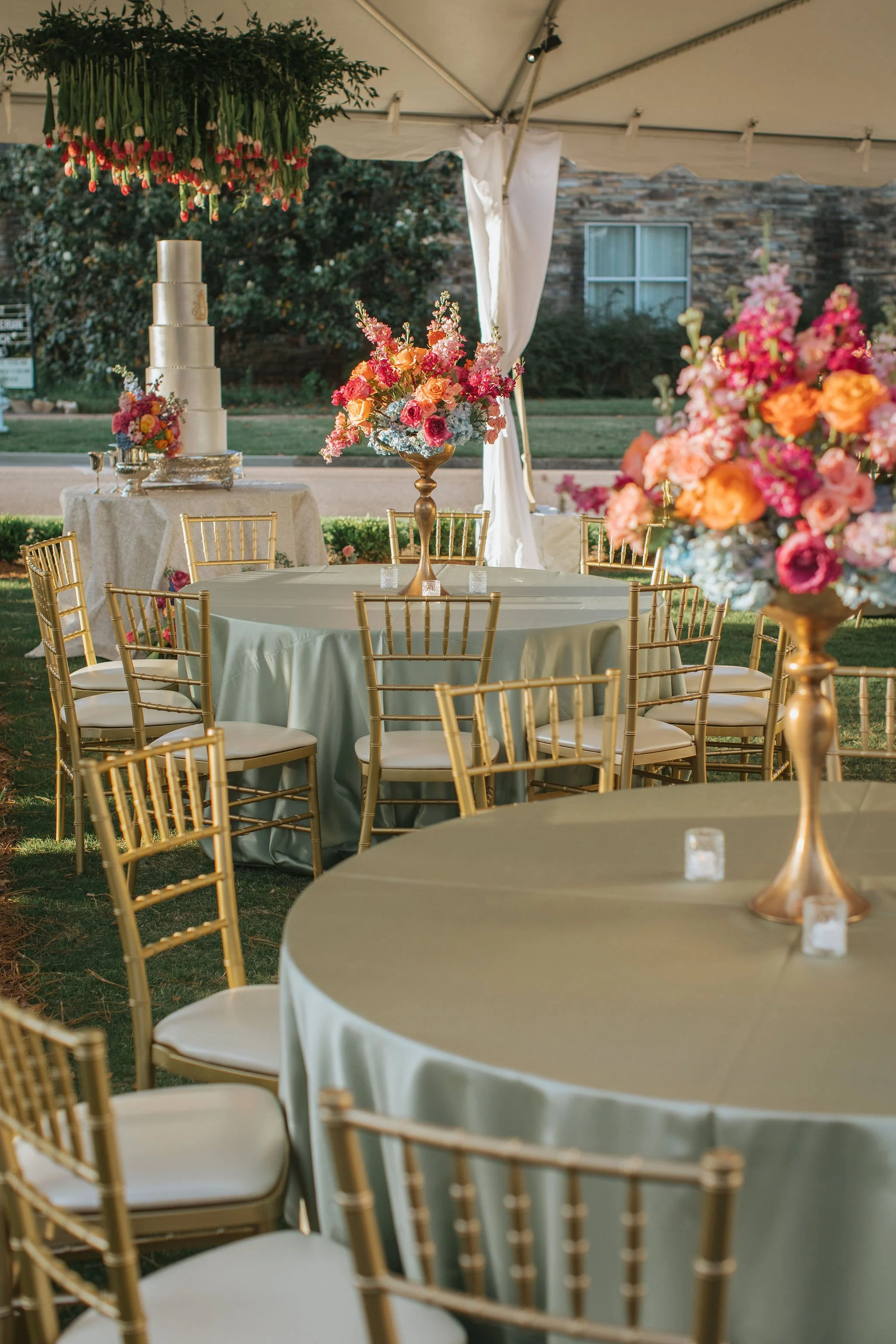 Elegant outdoor wedding set-up with round tables covered in light-colored tablecloths, gold chairs, floral centerpieces with pink, orange, and purple flowers, a multi-tiered white cake, and a tent with hanging floral arrangements.