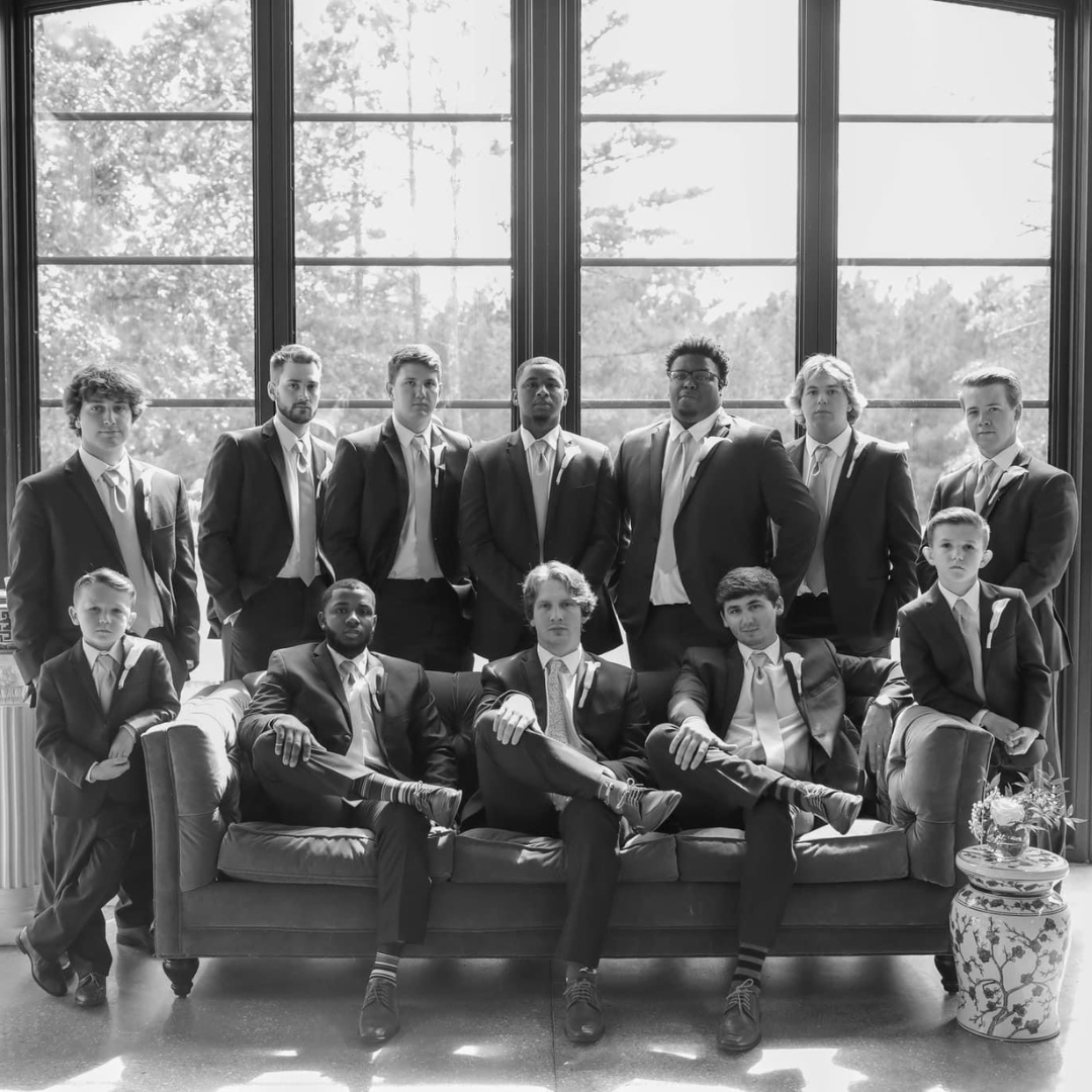 A group of twelve young men in suits posing for a photo in a large room with tall windows and trees outside.