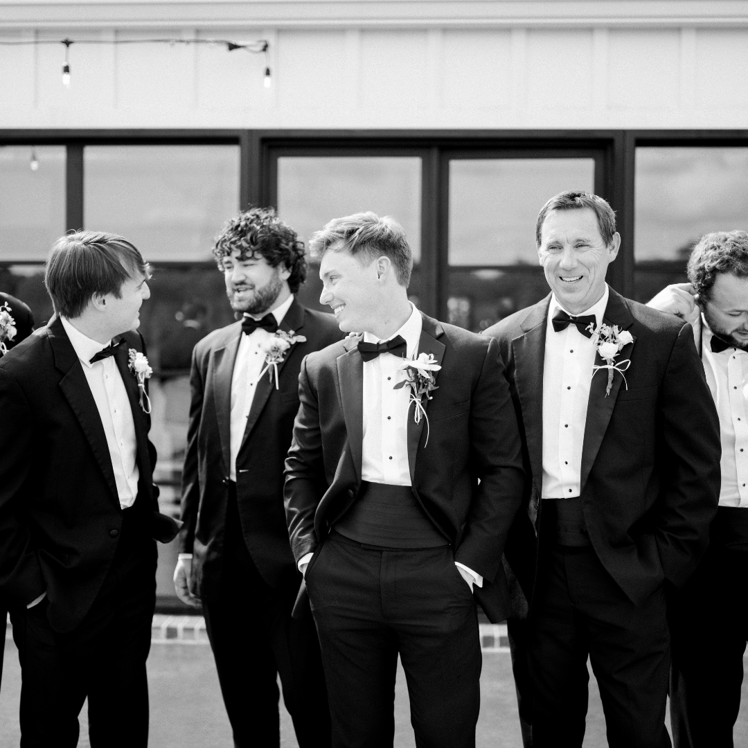 Group of men in tuxedos and bow ties standing and talking outside, smiling, with a building and windows in the background.