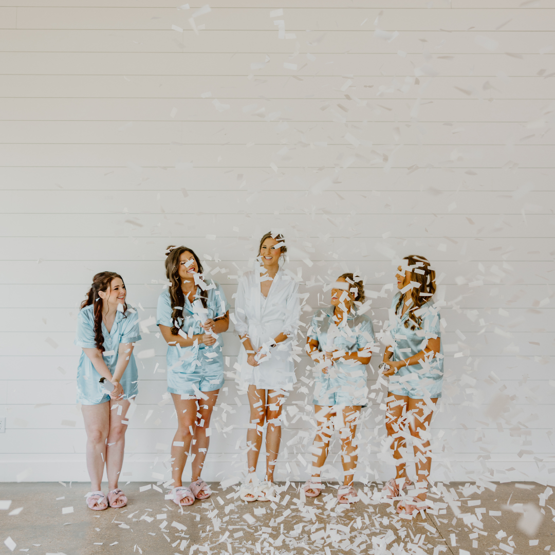 Group of five women in pajamas celebrating with confetti against a white wooden wall.