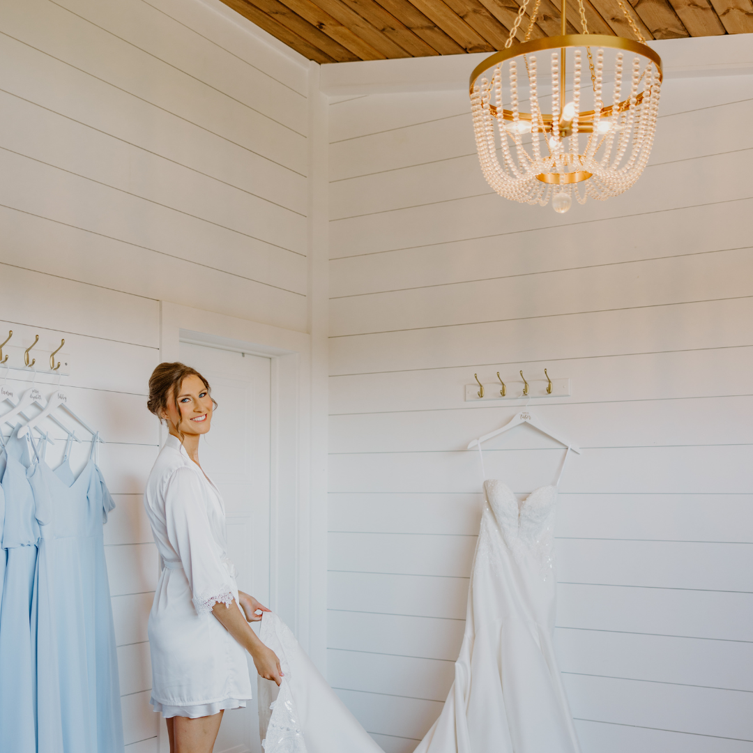 A woman in a white robe smiling while holding a wedding dress in a white room with a chandelier, a white door, and hanging gowns.