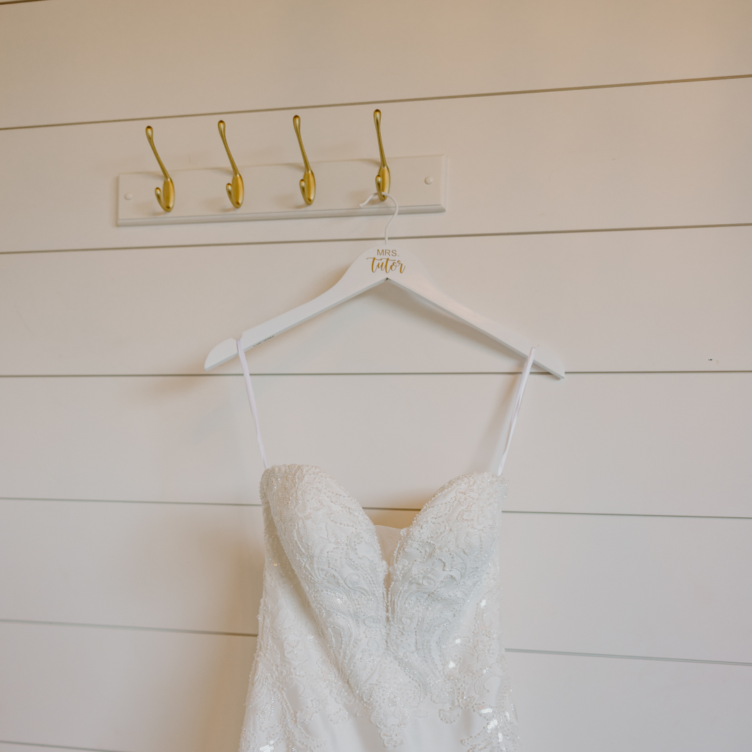 A white wedding dress with lace details hanging on a white hanger labeled 'Mrs. Tuter' on a wall with horizontal white wooden panels.