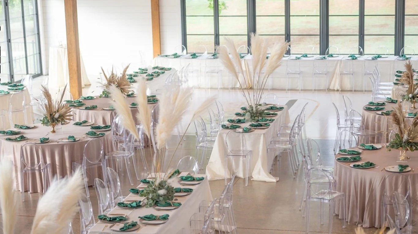 Elegant event space with round and rectangular tables decorated with greenery and pampas grass centerpieces, transparent chairs, large windows, and soft lighting.
