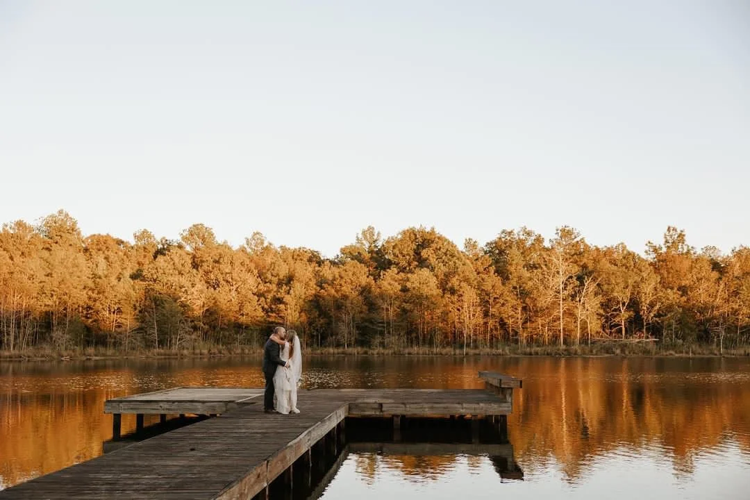 A couple in wedding attire stands on a wooden dock by a calm lake, embracing with trees in autumn colors in the background.