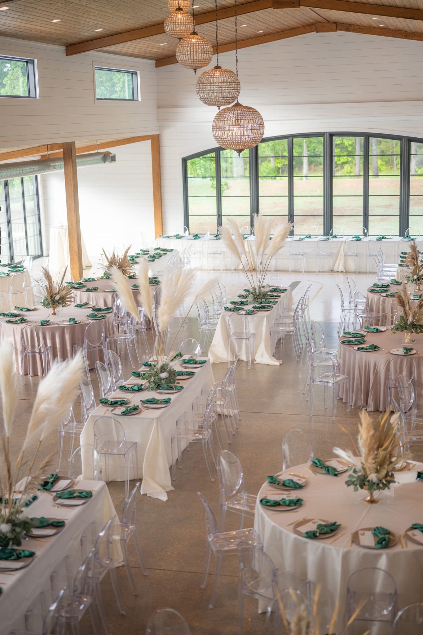 A decorated event or wedding reception hall with round and rectangular tables, green napkins, clear chairs, floral centerpieces with pampas grass, and wooden ceiling with hanging wicker light fixtures, large windows showing outdoor greenery.