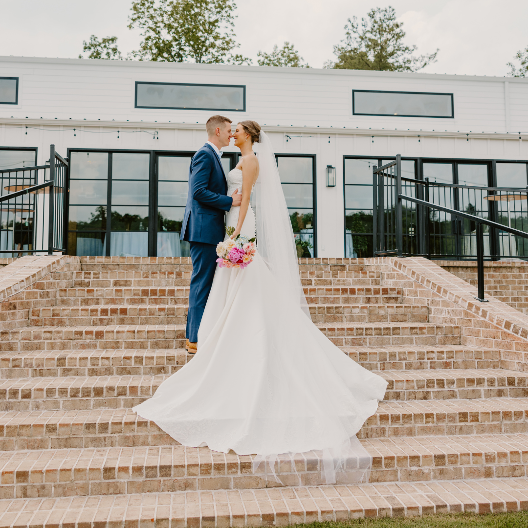 A bride and groom stand close together on brick steps outside a modern building, gazing into each other's eyes. The bride is wearing a white wedding gown and holding a bouquet of pink and white flowers, while the groom is dressed in a blue suit.