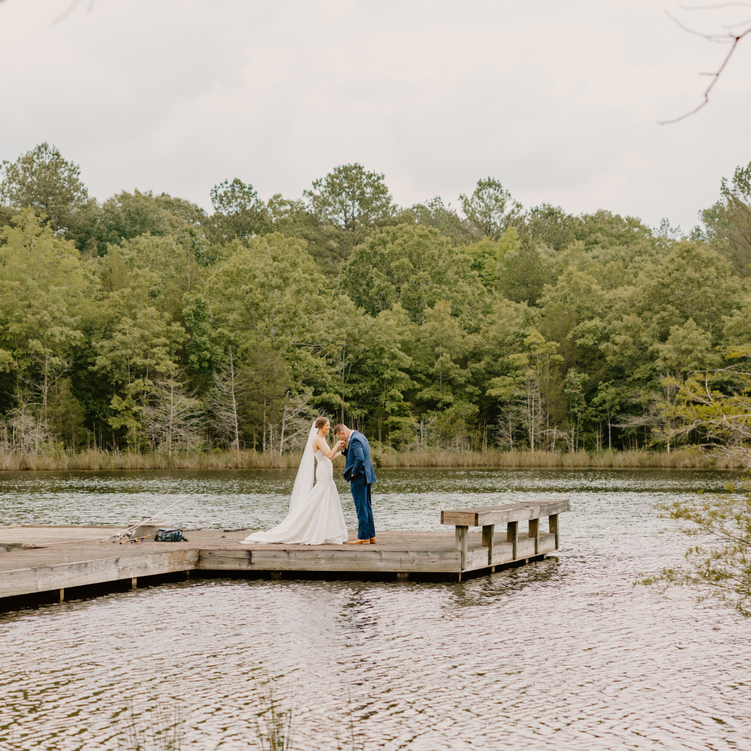 A bride and groom standing on a wooden dock by a lake, holding hands and kissing, with a background of green trees and an overcast sky.