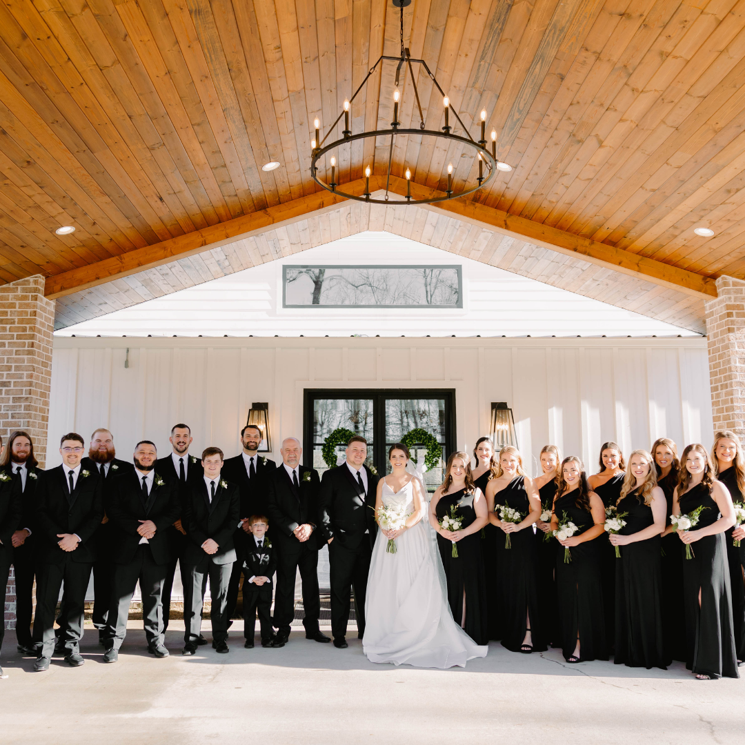 A wedding party standing in front of a white barn-style building with a wooden ceiling and a large chandelier, including the bride, groom, bridesmaids, groomsmen, and a young boy, all dressed in formal attire.