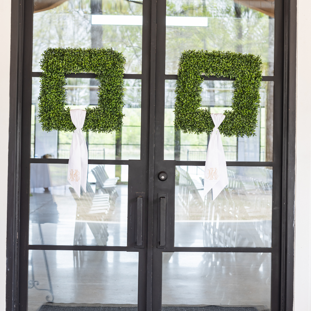 Decorative door with two square green wreaths adorned with white ribbons hung on a glass door, leading to an outdoor patio with chairs and trees visible in the background.