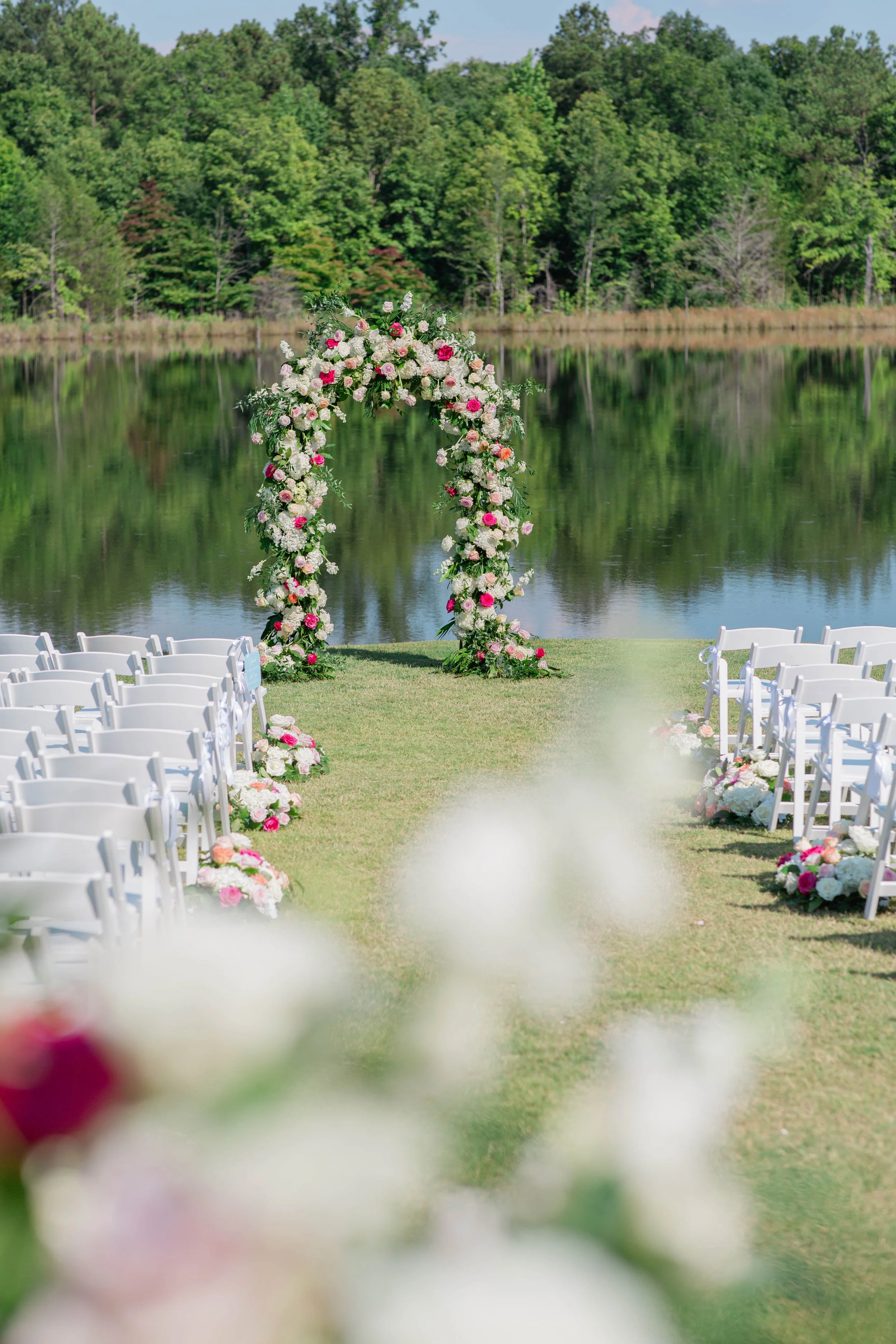 An outdoor wedding setup by a lake features white chairs arranged in rows on a grassy lawn. At the end of the aisle, there is a floral arch decorated with pink and white flowers, overlooking the water with green trees in the background.