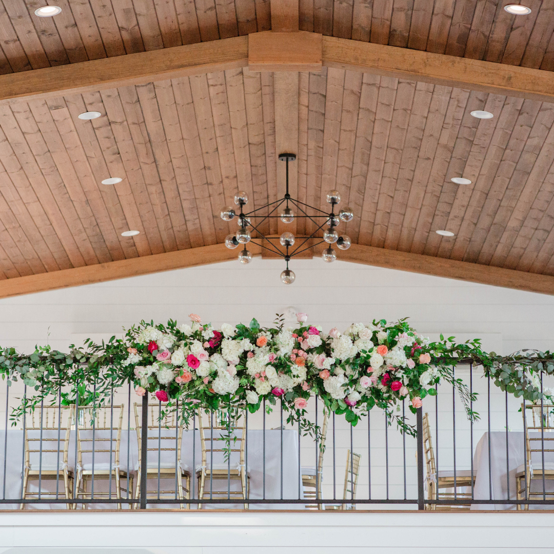 Interior of a decorated event space with floral arrangements on a railing, wooden ceiling, and a chandelier.