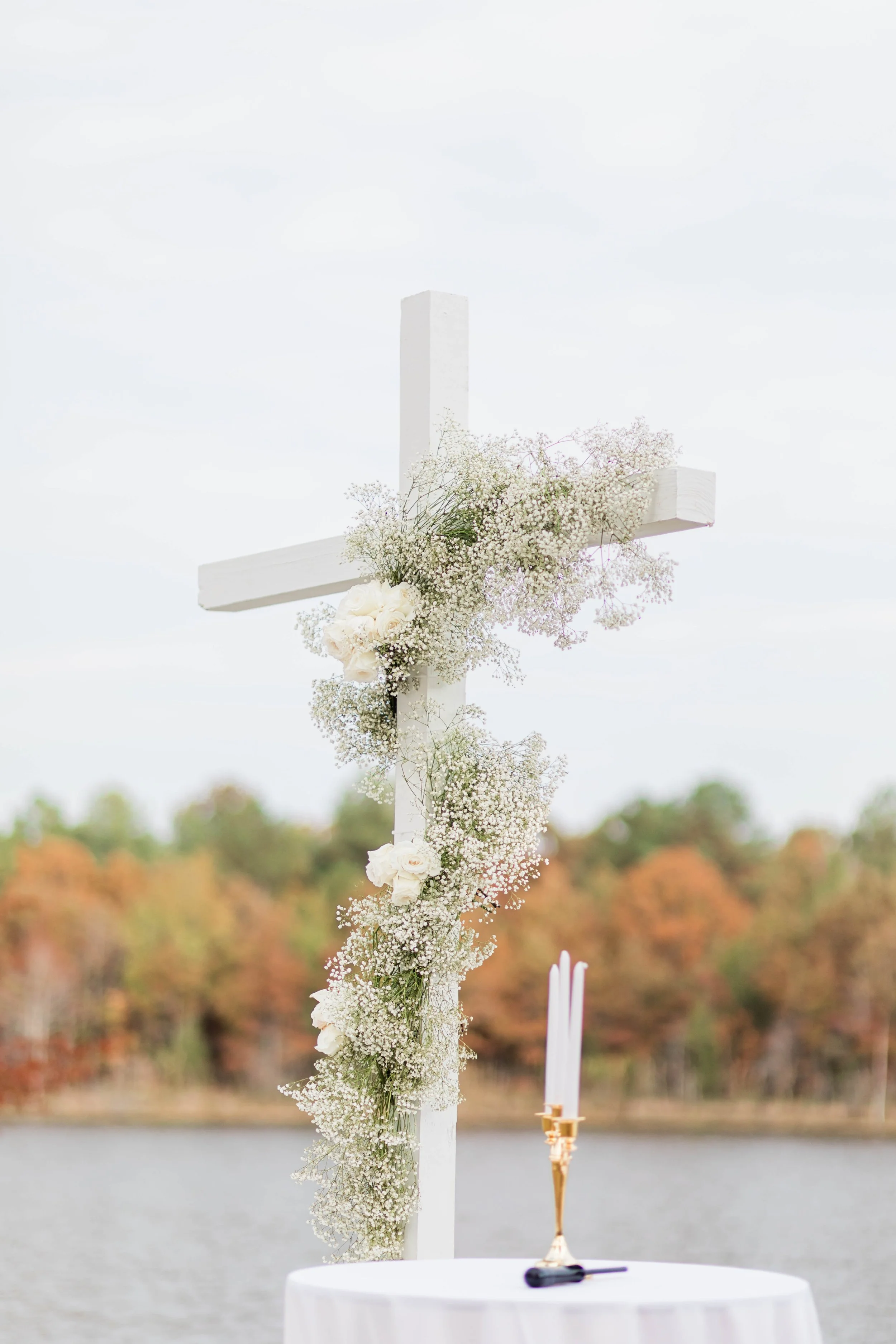 White wooden cross decorated with white roses and baby's breath flowers, set outdoors near a lake with autumn-colored trees in the background, with a table holding three white candles and a black lighter in the foreground.