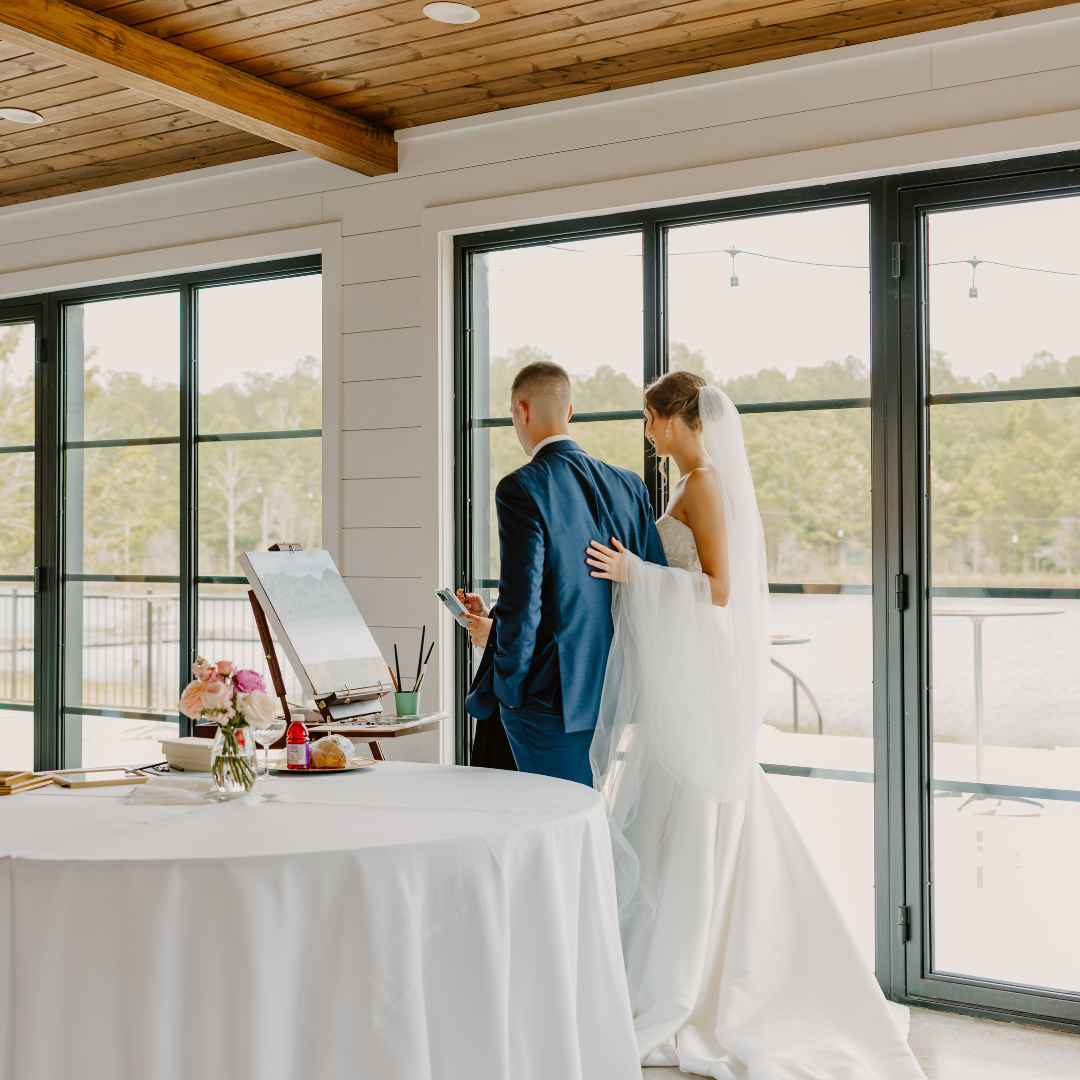 Bride and groom at their wedding ceremony, standing inside a room with large glass doors leading outside, with painting supplies and a table with flowers in the foreground.