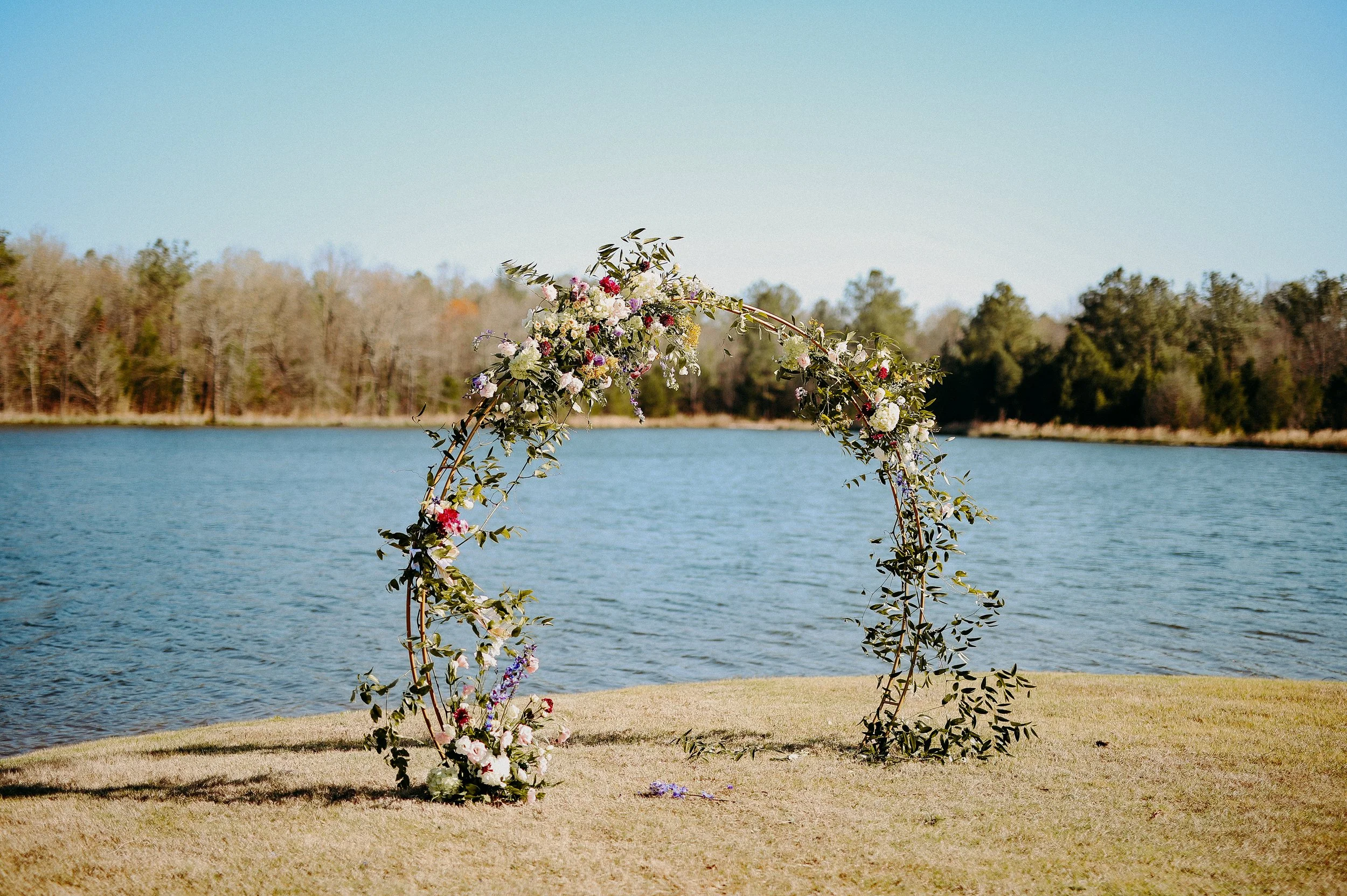 Floral wedding arch on grassy lakeshore with trees and water in the background.