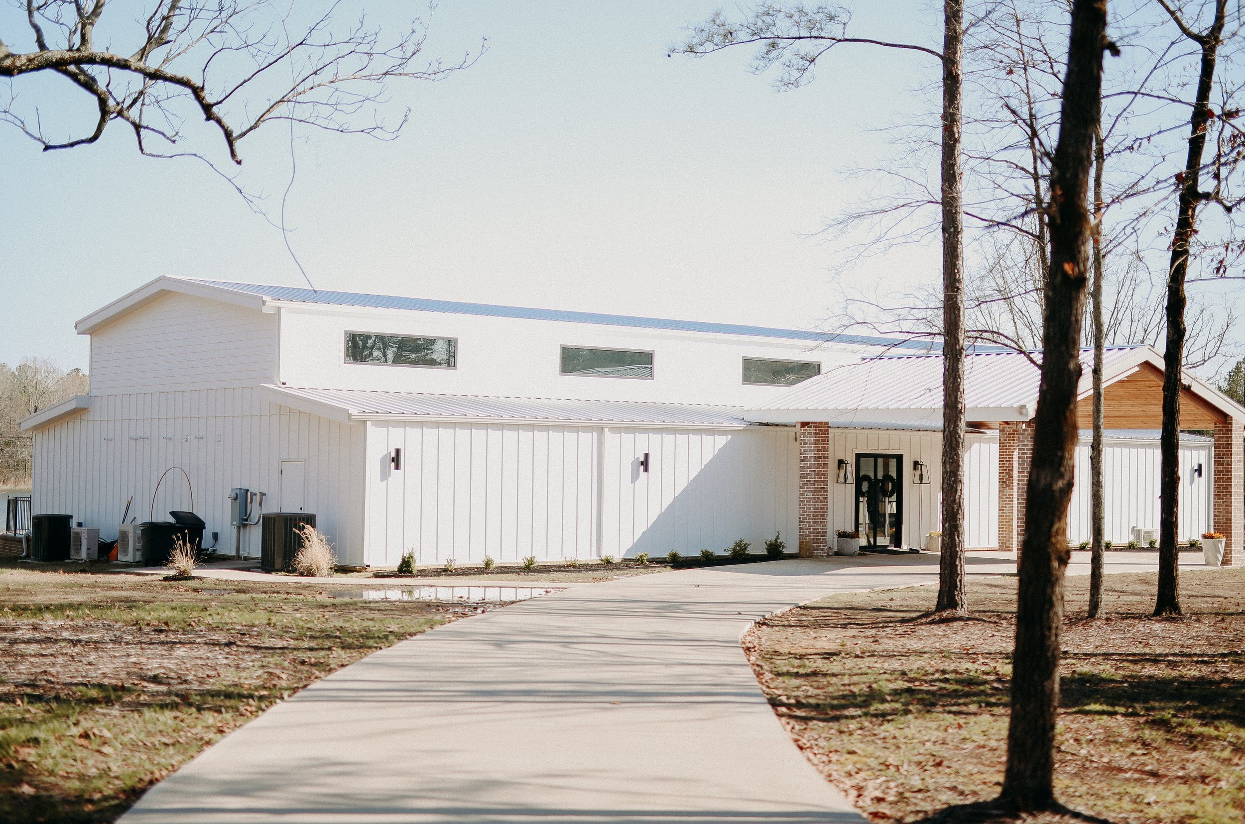 A modern white building with a metal roof, brick accents near the entrance, surrounded by leafless trees and a curved concrete pathway leading to the front door.