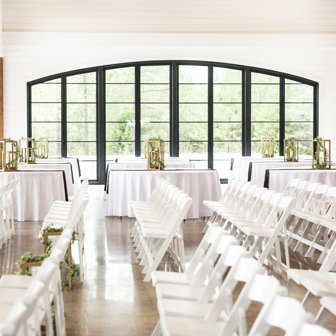 Set up interior for wedding reception with white folding chairs, round tables with white tablecloths, and decorative lantern centerpieces, large arched window overlooking greenery.