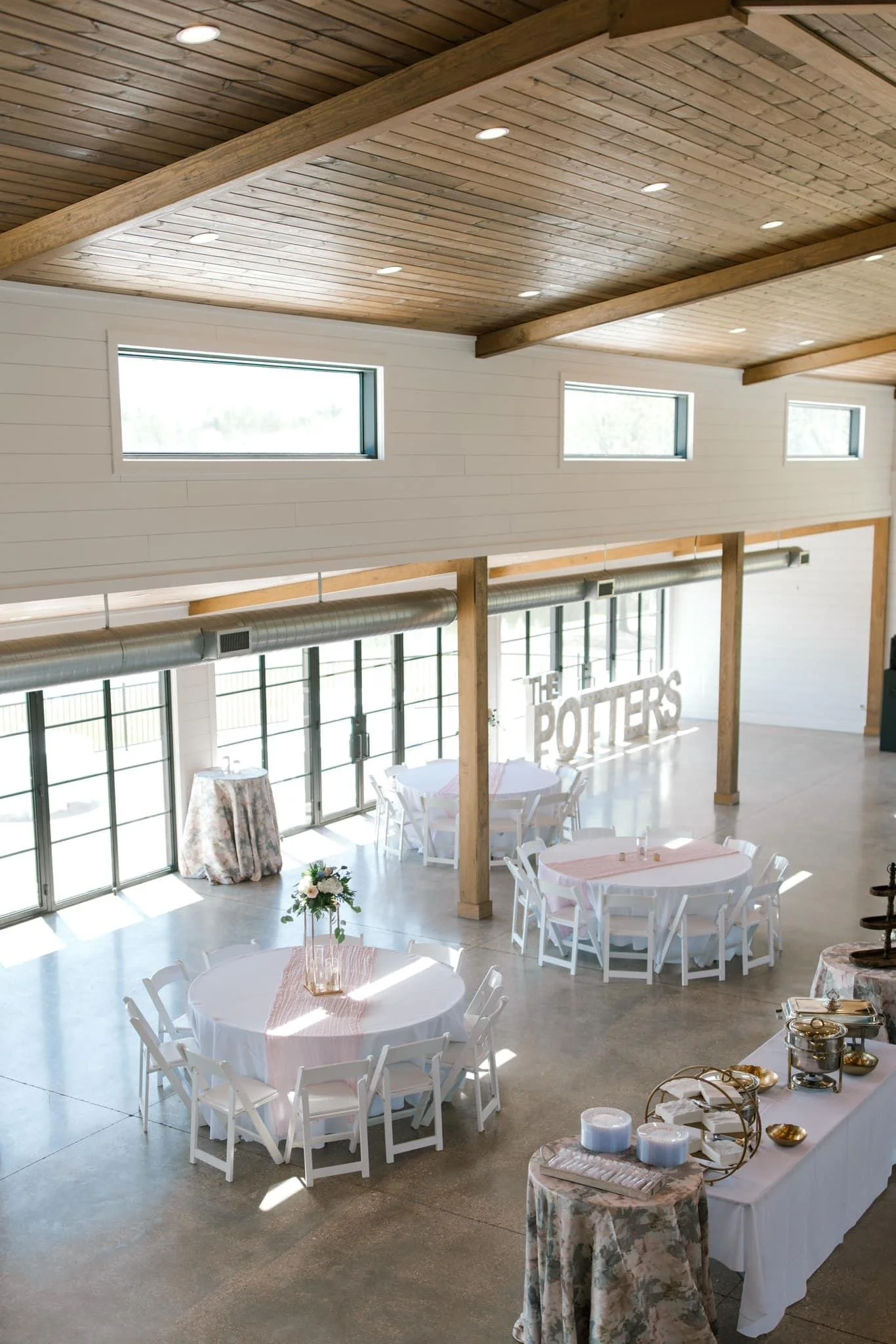 Interior of a spacious event venue with high wooden ceilings, multiple rectangular windows, round tables with white chairs, and a small table with decorative items. Large sign reading 'The Poeters' is visible in the background.