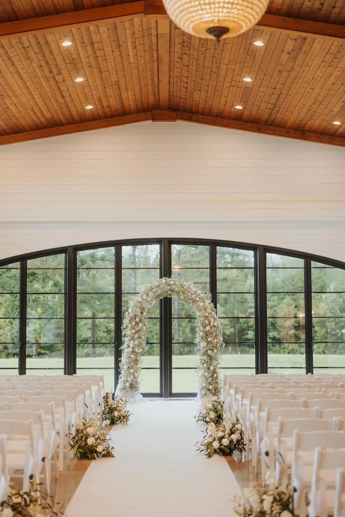 Indoor wedding ceremony setup with white floral arch over the aisle, beige chairs on both sides, large glass windows, high wooden ceiling, and wooden flooring.