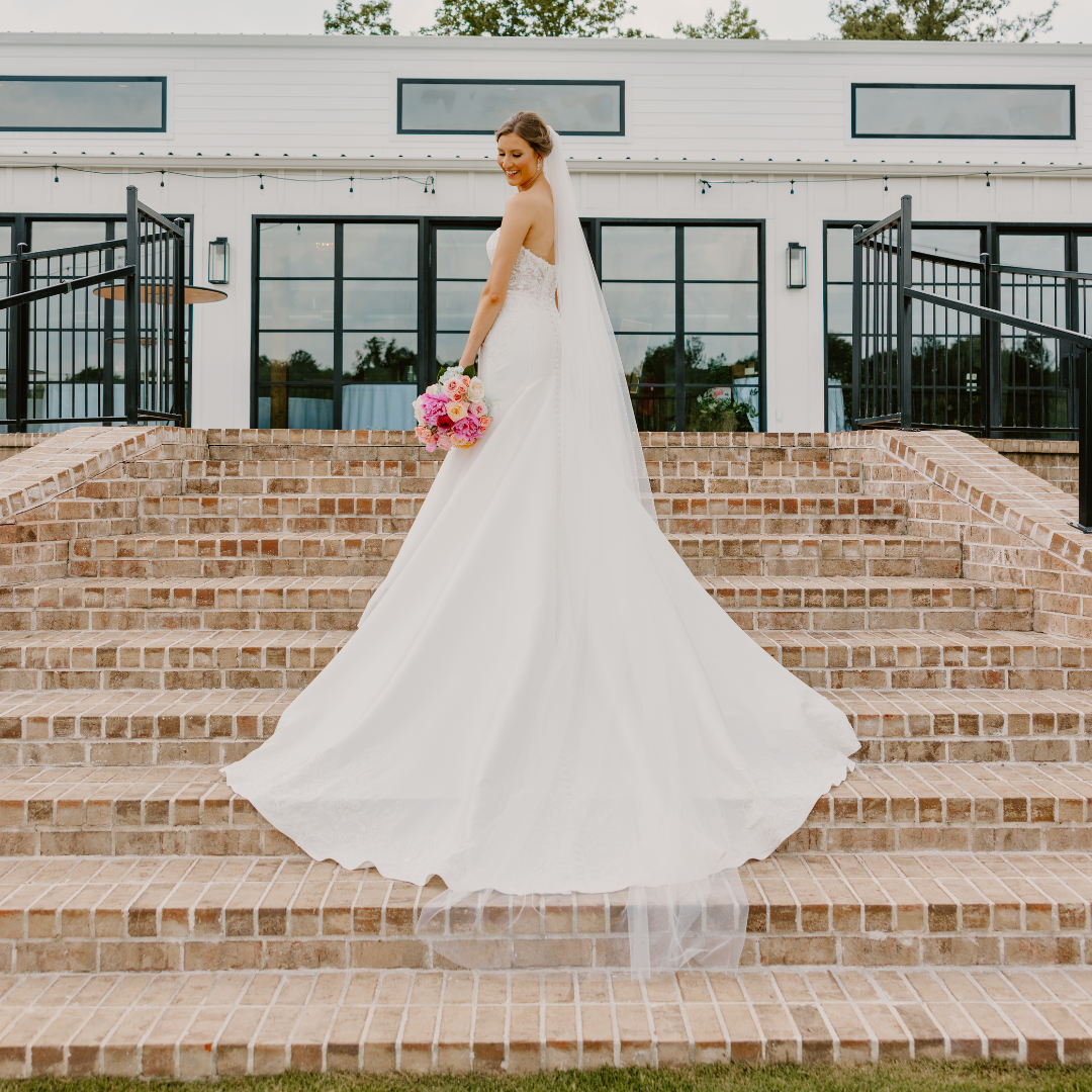 Bride in white wedding dress holding pink and white bouquet, standing on brick steps outside building with large windows and black railings.