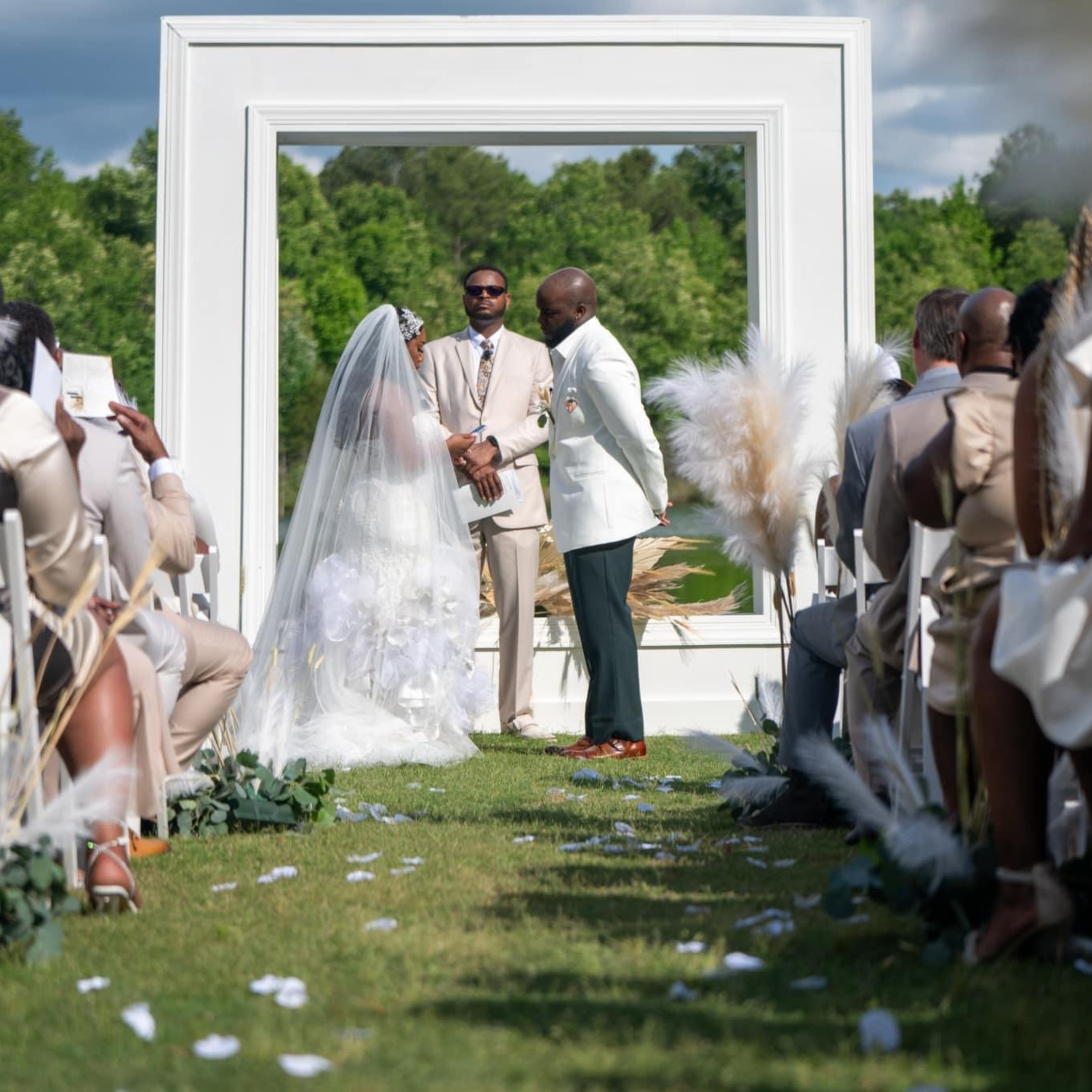 A wedding ceremony outdoors with a bride, groom, and officiant standing in front of a white frame with green trees and blue sky in the background. Guests are seated on either side.