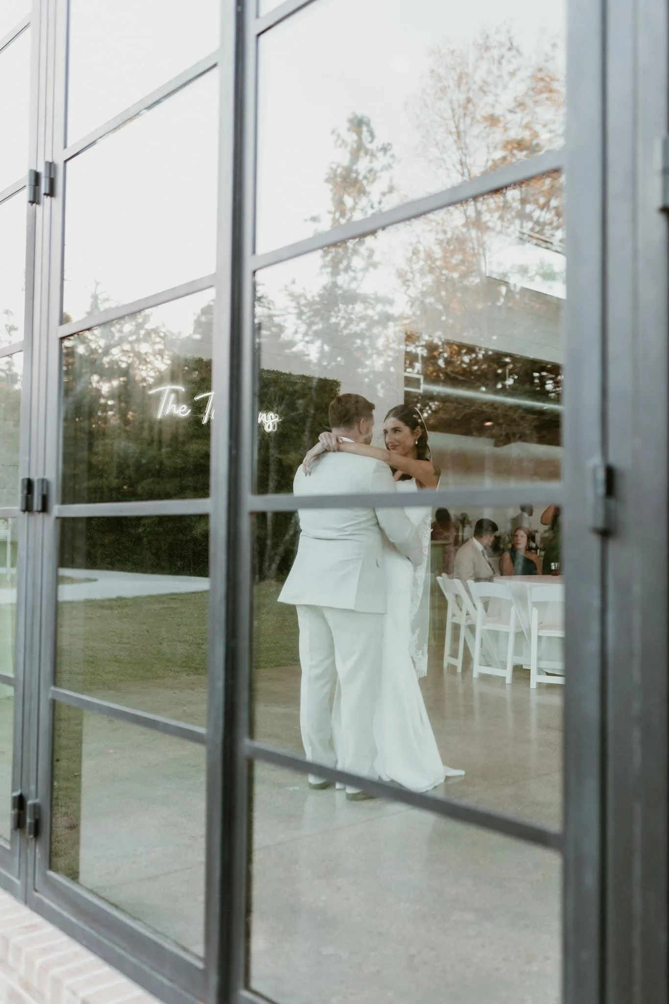 A bride and groom dressed in white are dancing during their wedding reception, viewed through a glass window.