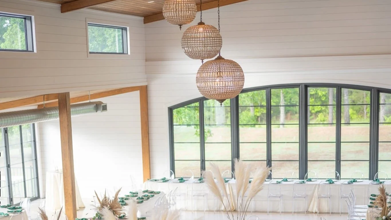 Interior of a bright, modern event space with large arched windows, hanging woven pendant lights, and tables set with plates and napkins, decorated with pampas grass.
