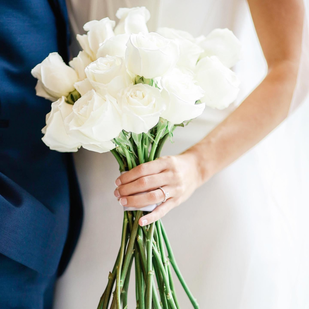 A bride holding a bouquet of white roses with her left hand, with a wedding ring visible.