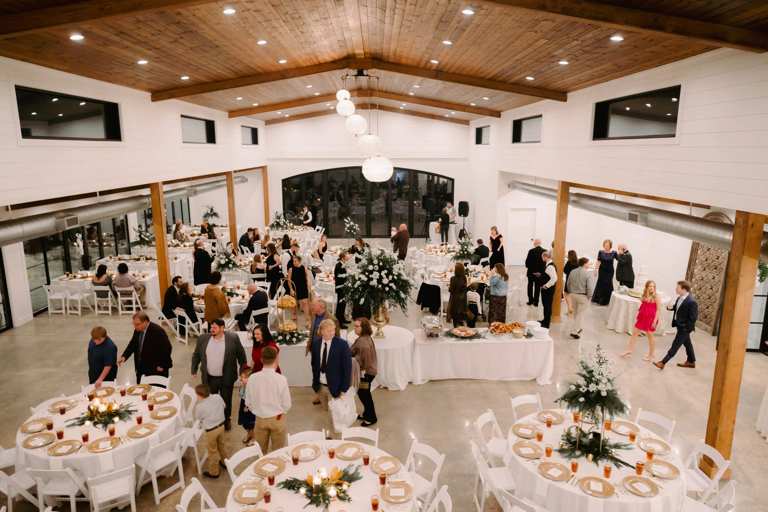 A large banquet hall with wooden ceiling and white walls decorated for a celebration. Guests are mingling and seated at round tables with white tablecloths and floral centerpieces, and there is a buffet table with food.