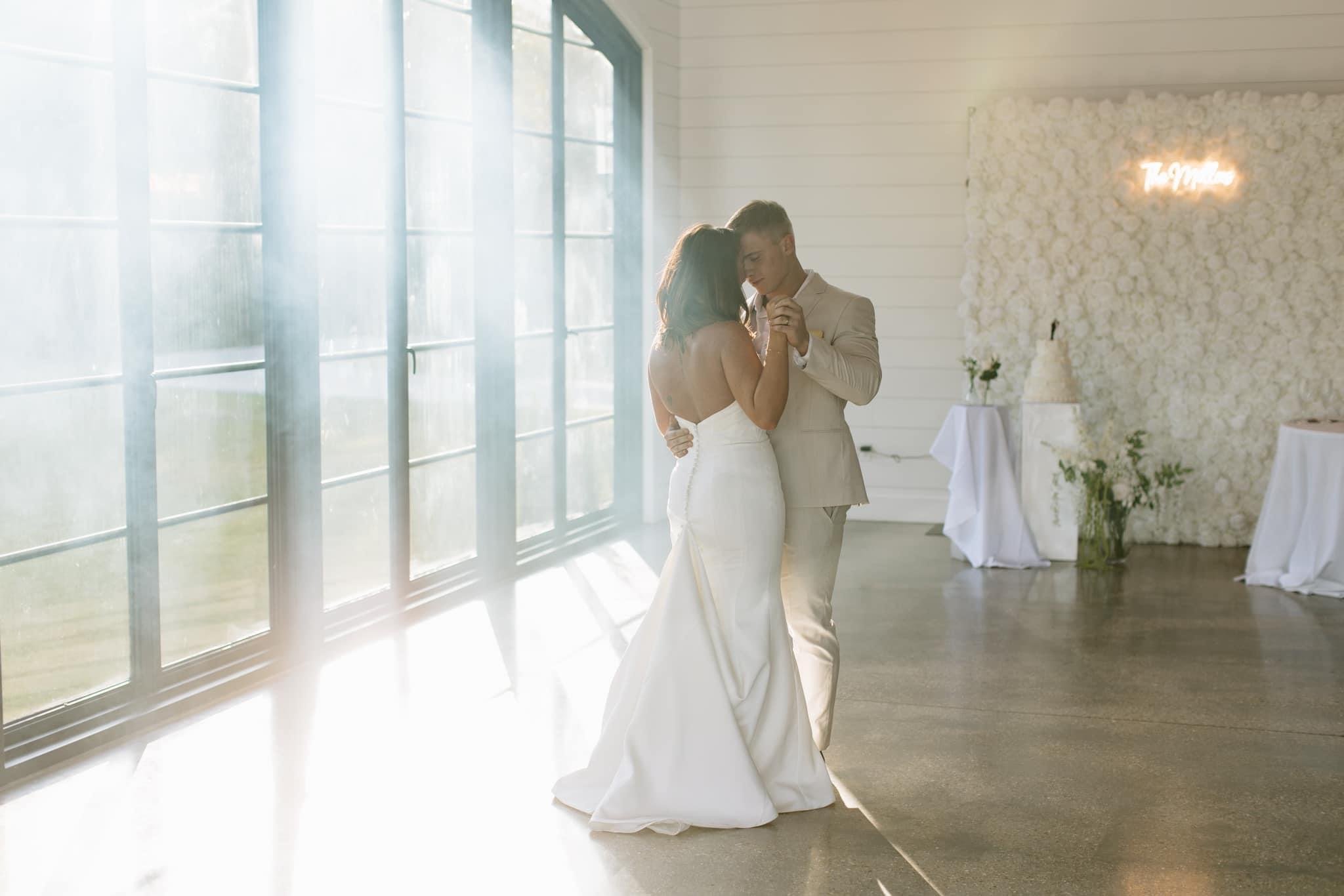 A bride and groom dancing inside a bright wedding venue with large window panels and white floral decorations on the wall.