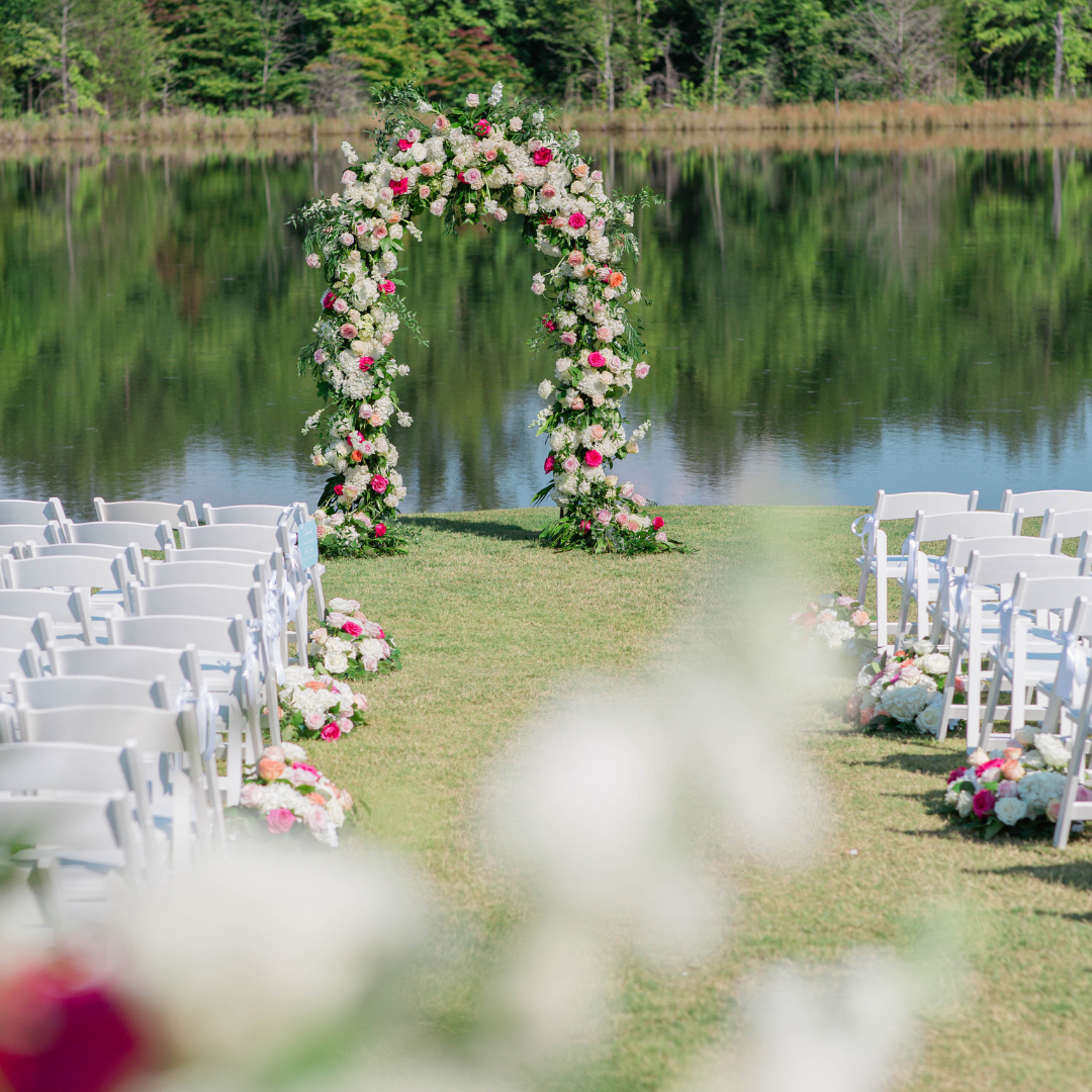Outdoor wedding altar decorated with a floral arch near a lake, with white chairs on either side and floral arrangements on the grass.