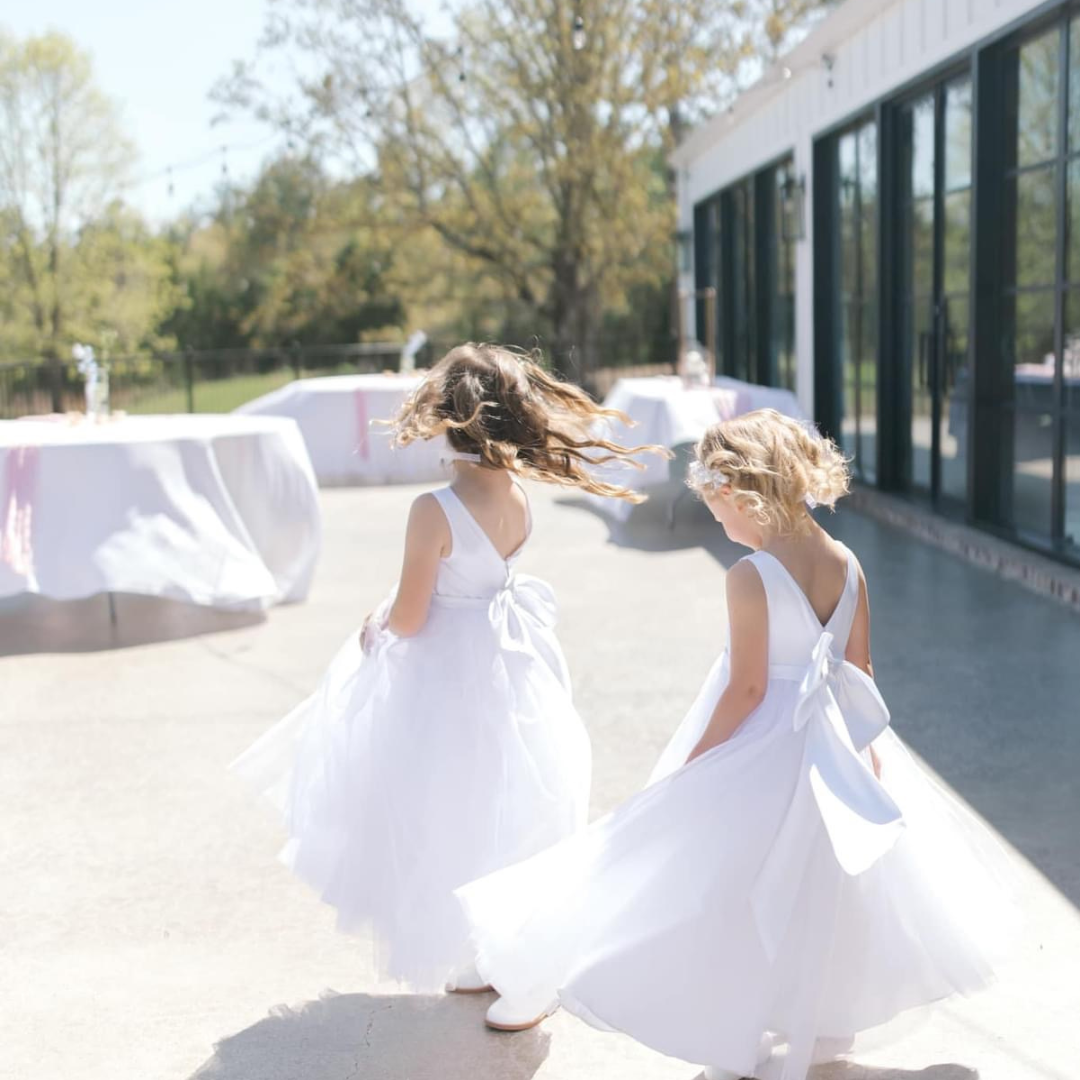 Two young girls in white dresses spinning and dancing outside during daytime at a celebration or wedding event, with tables set up in the background.