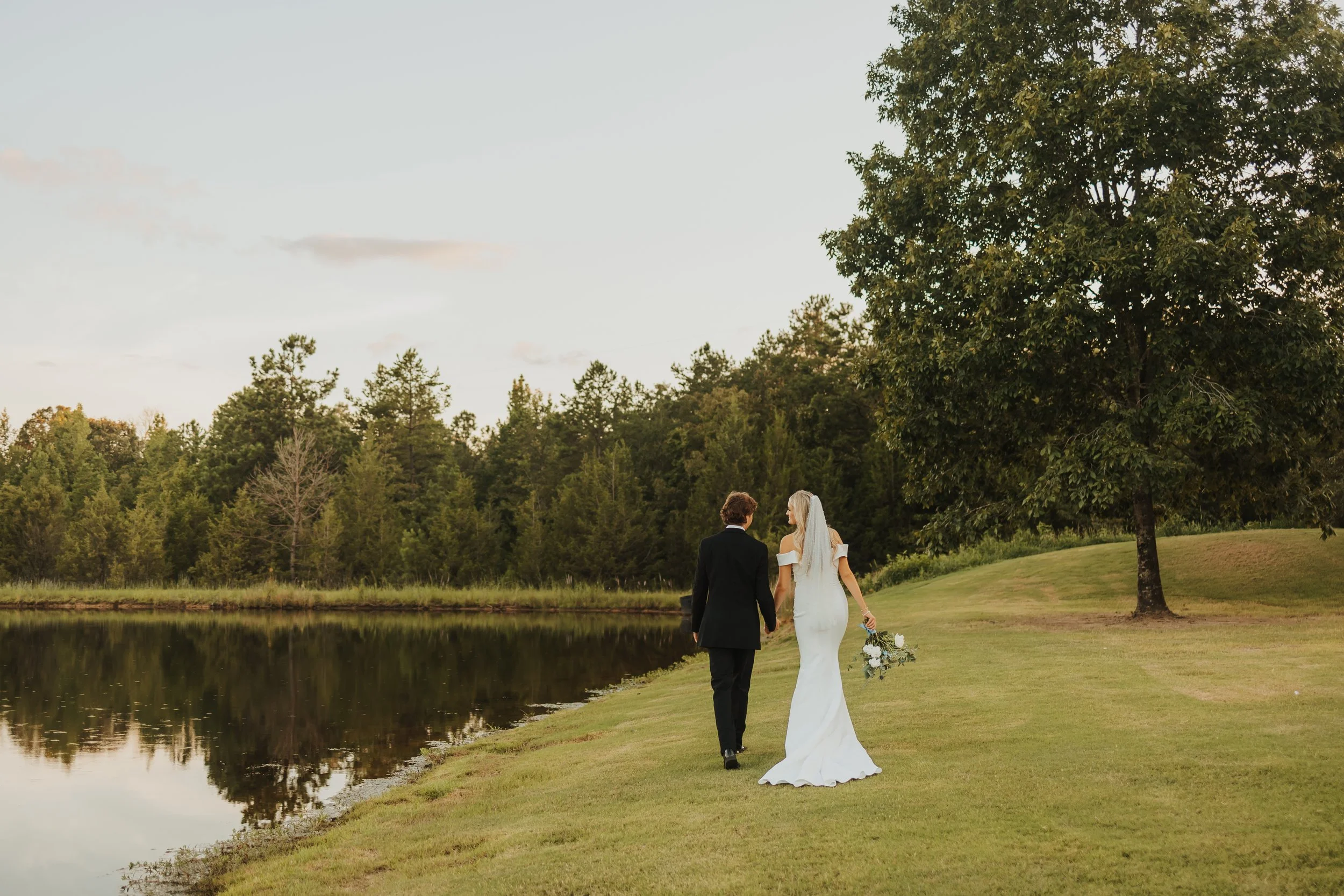 A bride and groom walking hand in hand by a lake in a wooded outdoor setting during sunset, with the bride holding a bouquet of flowers.