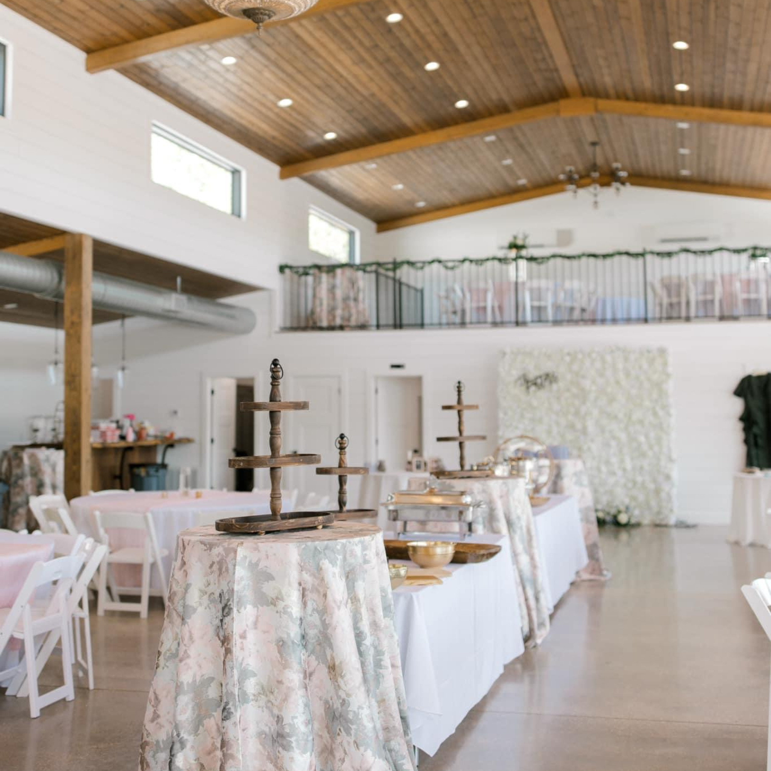 Interior of a decorated event space with tables covered in floral tablecloths, wooden cake stands, and buffet arrangements. The room has a wooden ceiling with recessed lighting, white walls, and an upper balcony with railing decorated with garland.