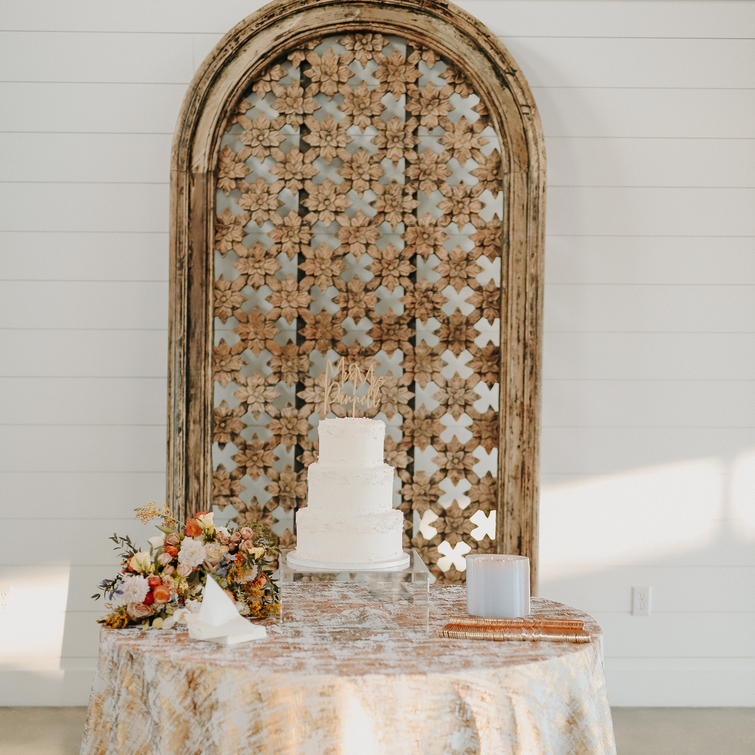 A three-tiered white wedding cake with a "Mama Dandelion" cake topper on a round table with a patterned tablecloth. Next to the cake is a bouquet of flowers and a white card. Behind the table is a decorative wooden panel with a floral pattern, attach