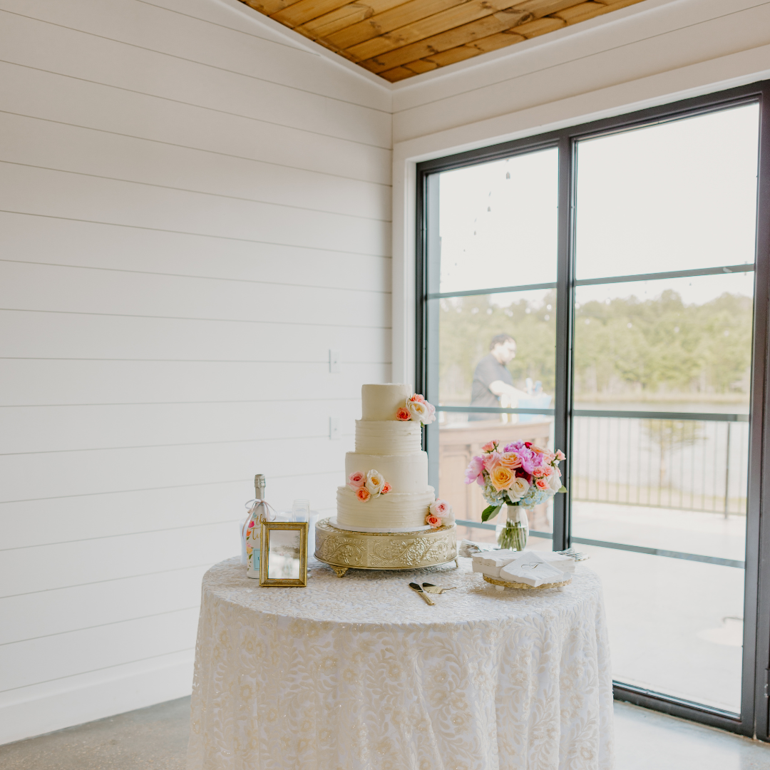 A three-tiered white wedding cake with pink and orange flowers, placed on a decorated table with a vase of pink and orange flowers, a bottle, a small picture frame, a cake knife and server, and napkins near a large window overlooking a scenic outdoor