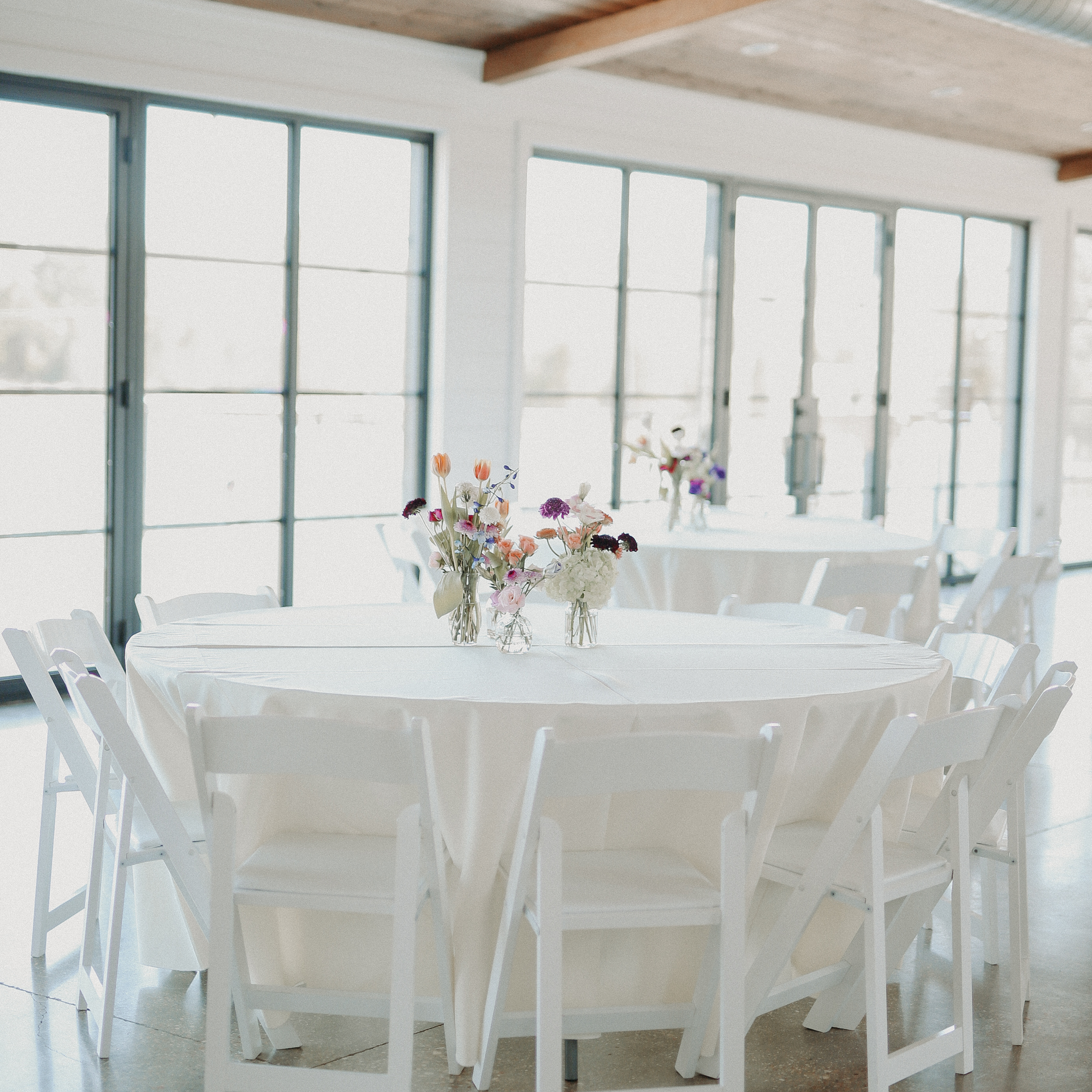 Round table with white tablecloth and floral centerpieces in a bright room with large windows.