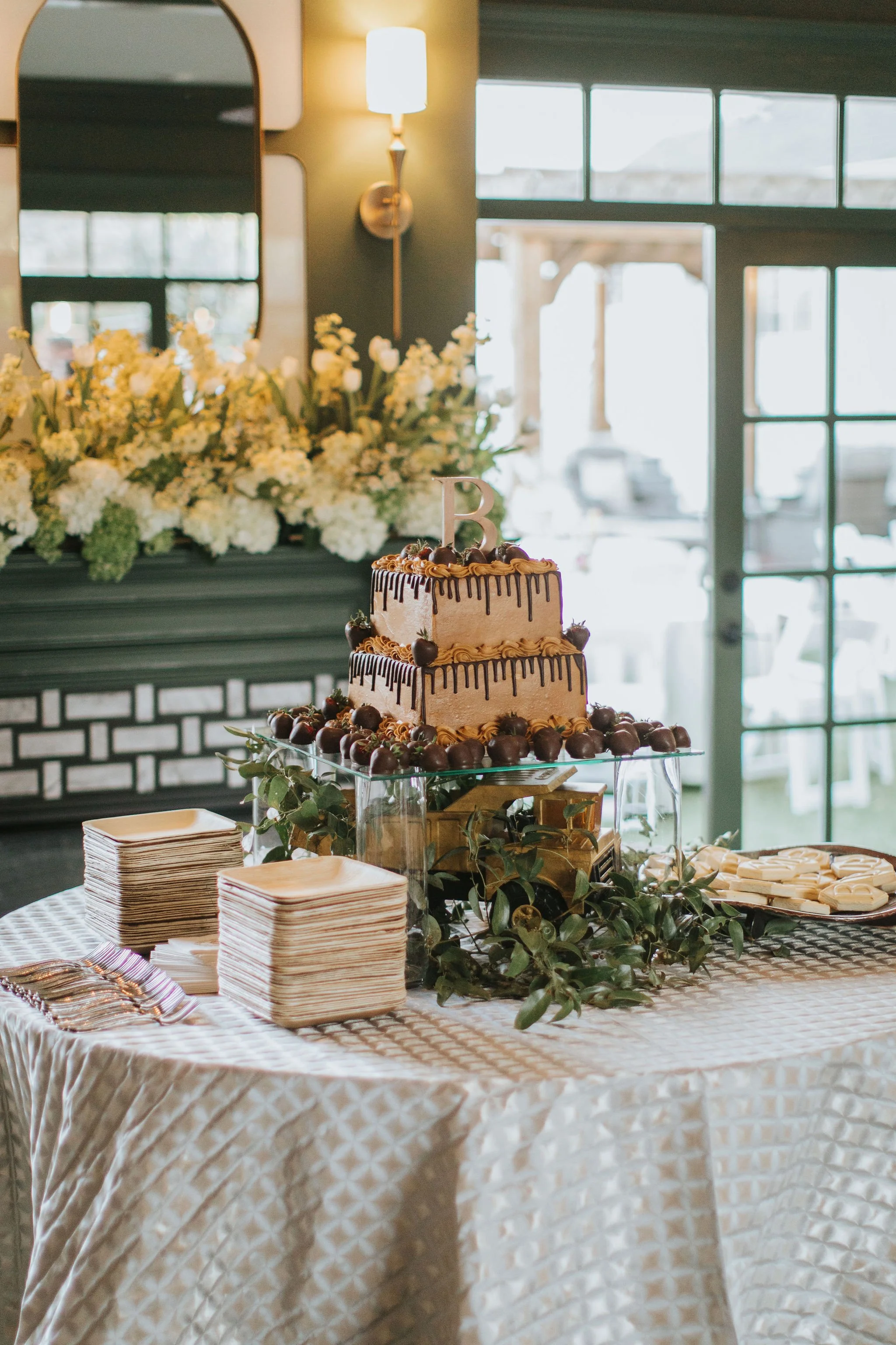 A decorated table with a cake, plates, napkins, and greenery, set for a celebration in a bright, airy room with large windows and floral arrangements in the background.