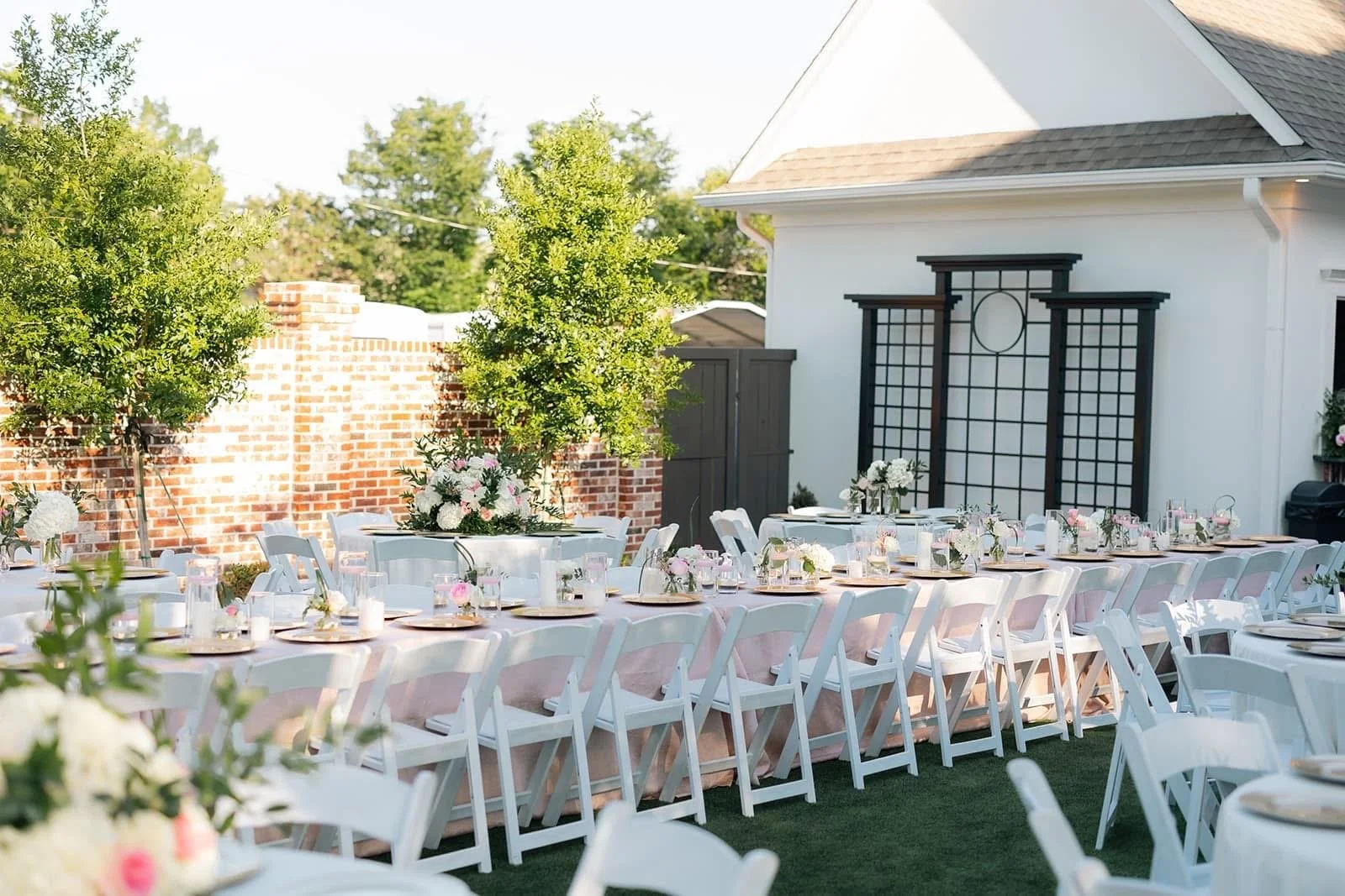 Outdoor reception decorated with long tables covered with pink tablecloths, surrounded by white chairs, and adorned with floral centerpieces and candles, set against a white house with black lattice panels and a brick wall in the background.